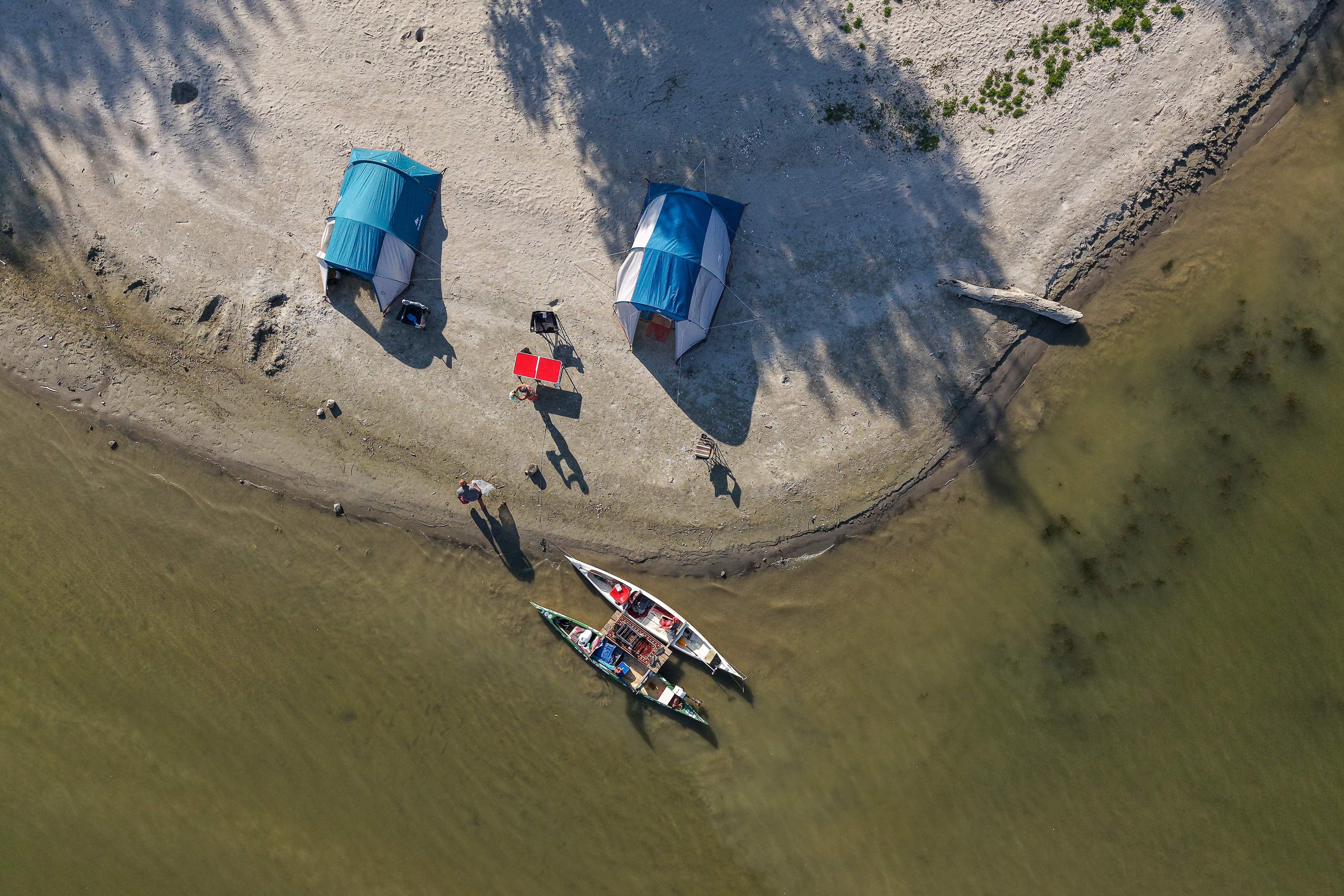 Zwei Zelte, Kanus und Menschen auf einer Sandbank am flachen Ufer der Donau aus der Vogelperspektive.