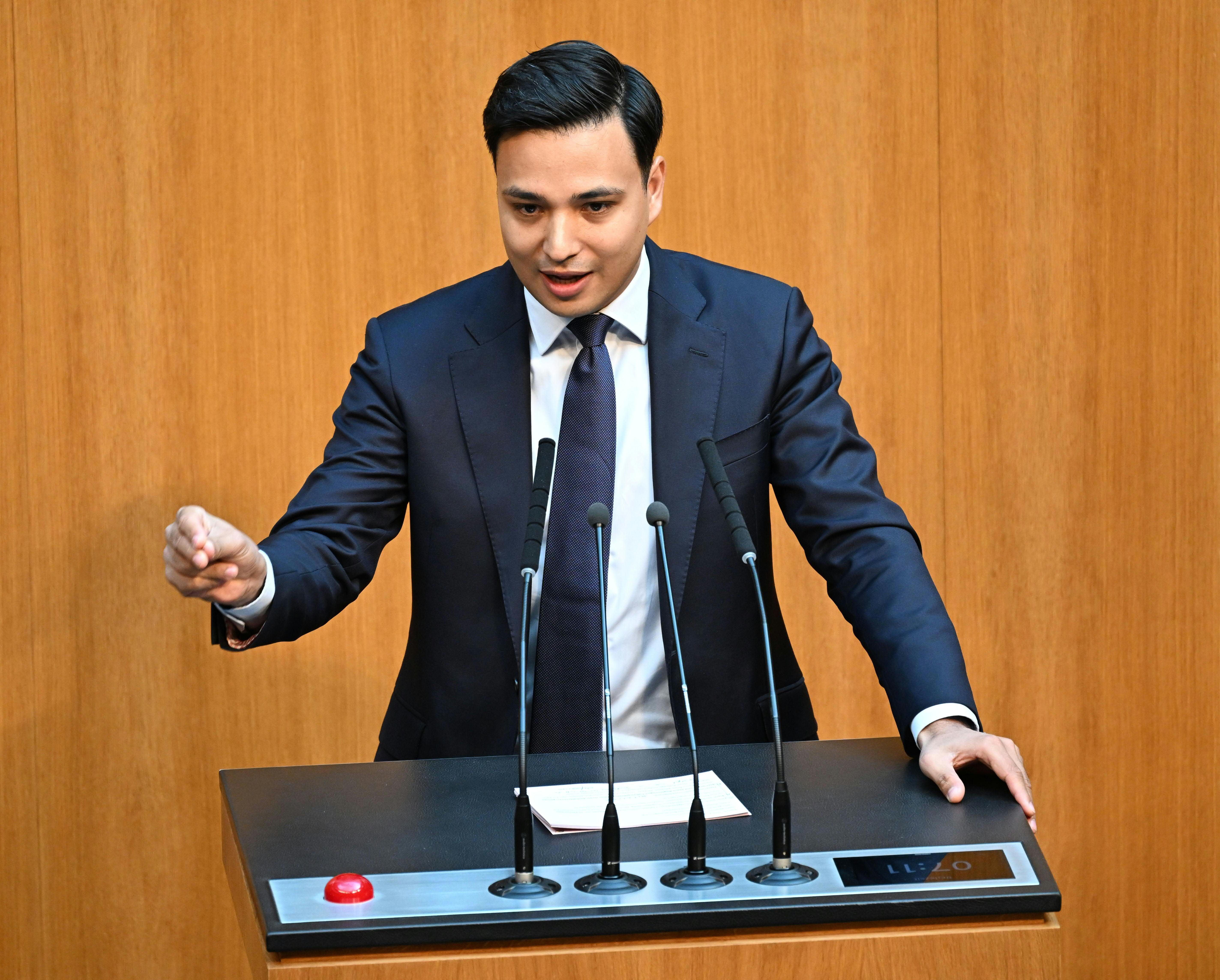 Ein Mann im Anzug spricht an einem Rednerpult mit Mikrofonen im Parlament.