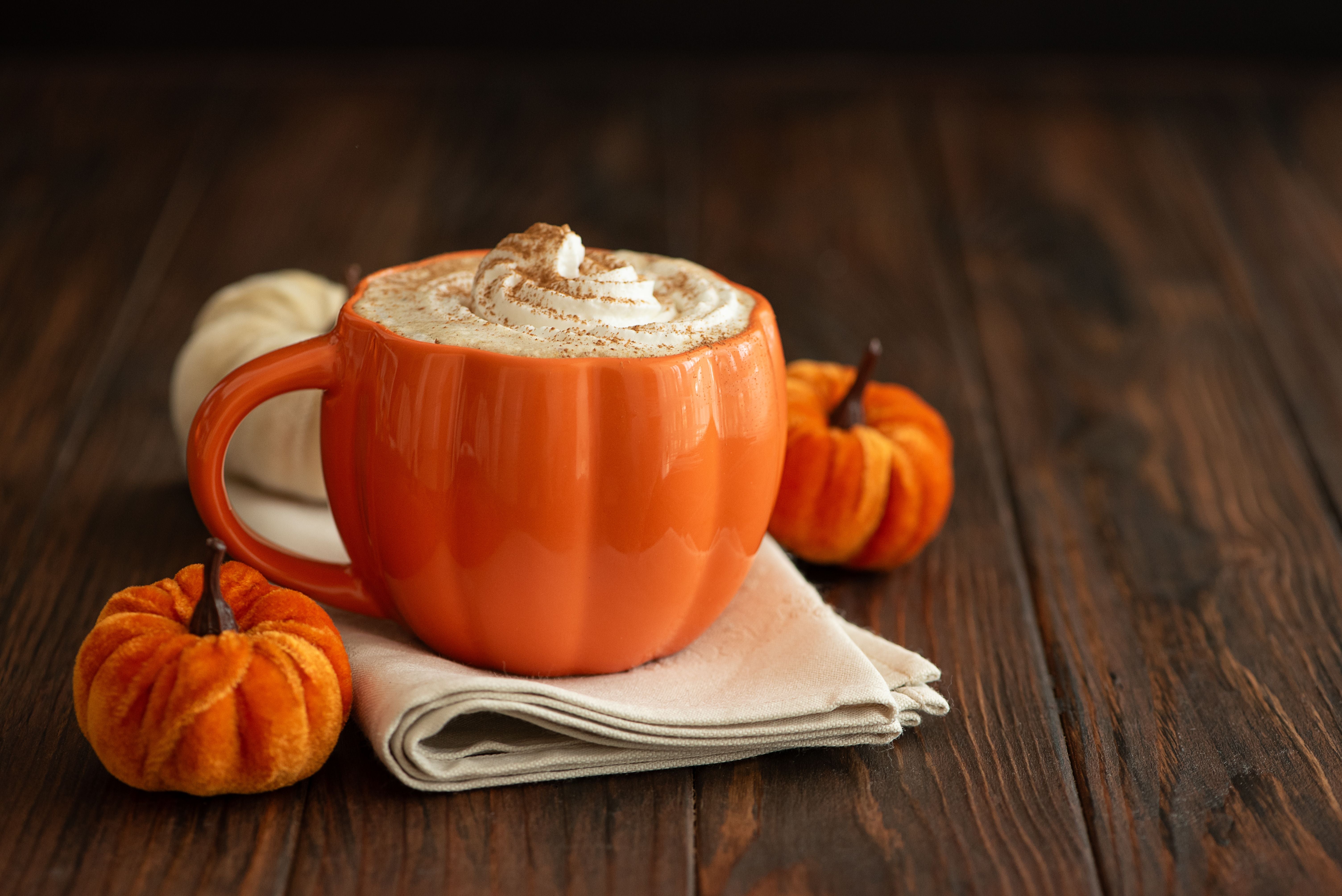 Kürbis-Latte in einer kürbisförmigen Tasse mit Sahne, umgeben von Herbstdeko auf Holz.