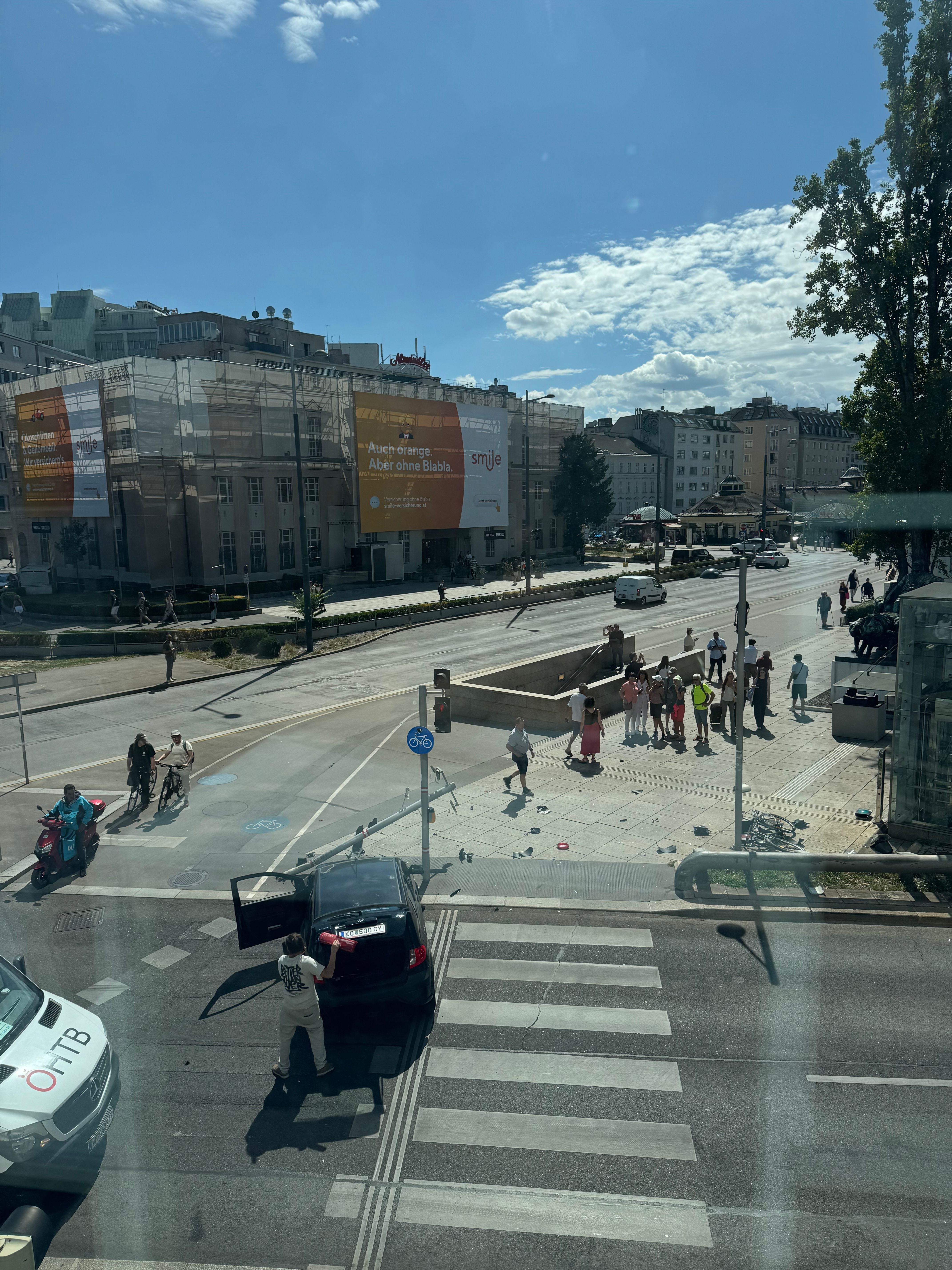Straßenszene mit Auto an der Ampel, Mann am Kofferraum und mehreren Fußgängern auf dem Gehweg.
