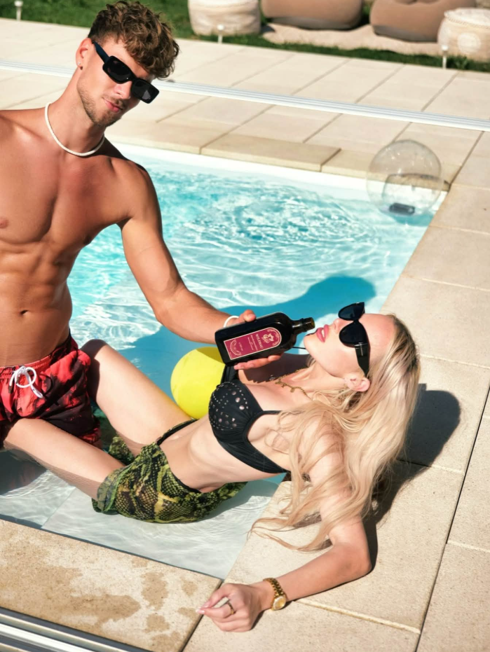 Mann reicht Frau am Pool eine Flasche, beide tragen Sonnenbrillen und Badesachen.