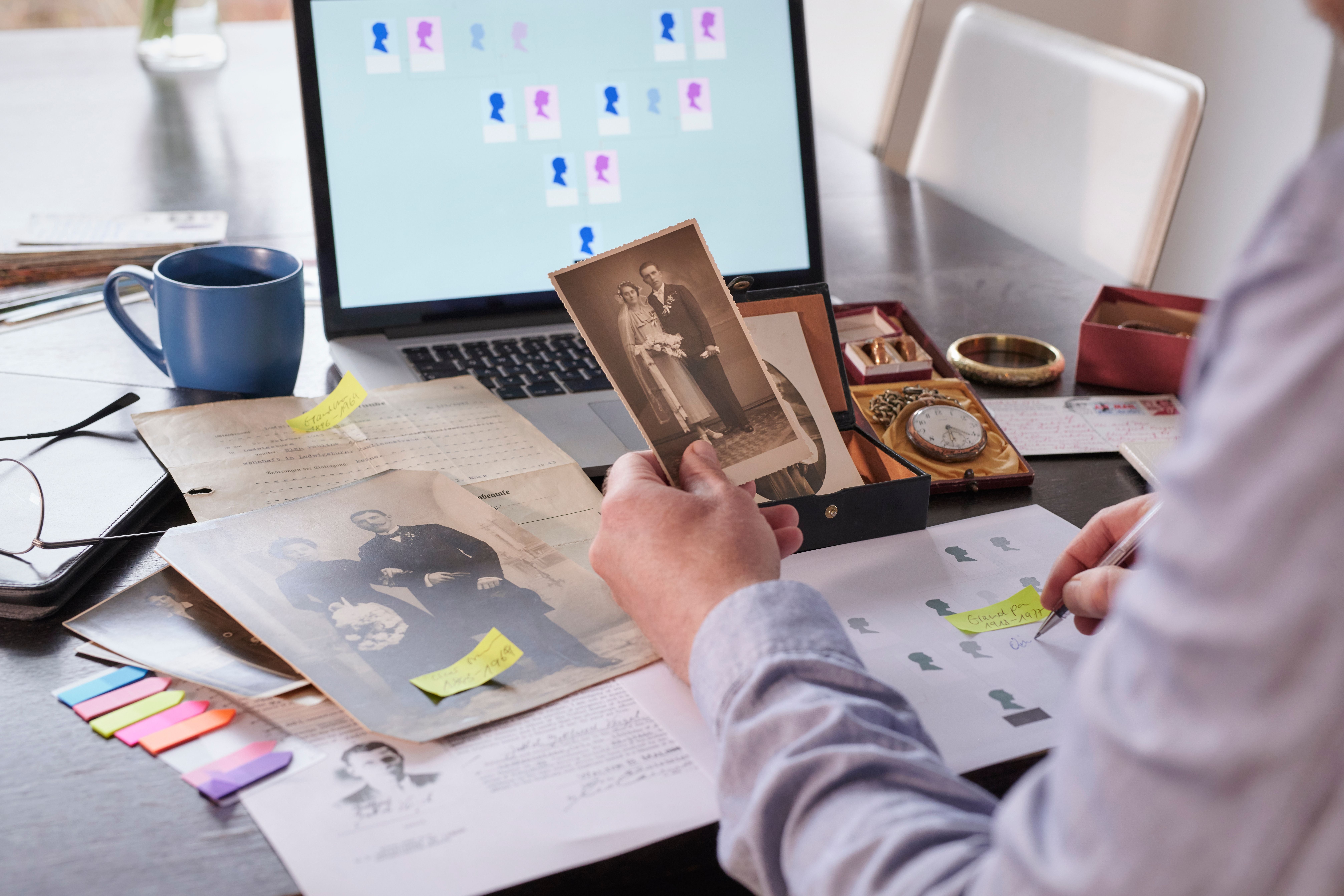 Mann recherchiert am Tisch mit alten Fotos und Laptop Ahnenforschung für einen Stammbaum.