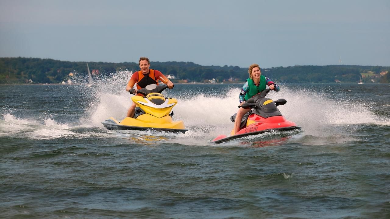 Zwei Personen fahren lachend auf Jetskis über das Wasser, einer gelb, einer rot, im Hintergrund Küste.
