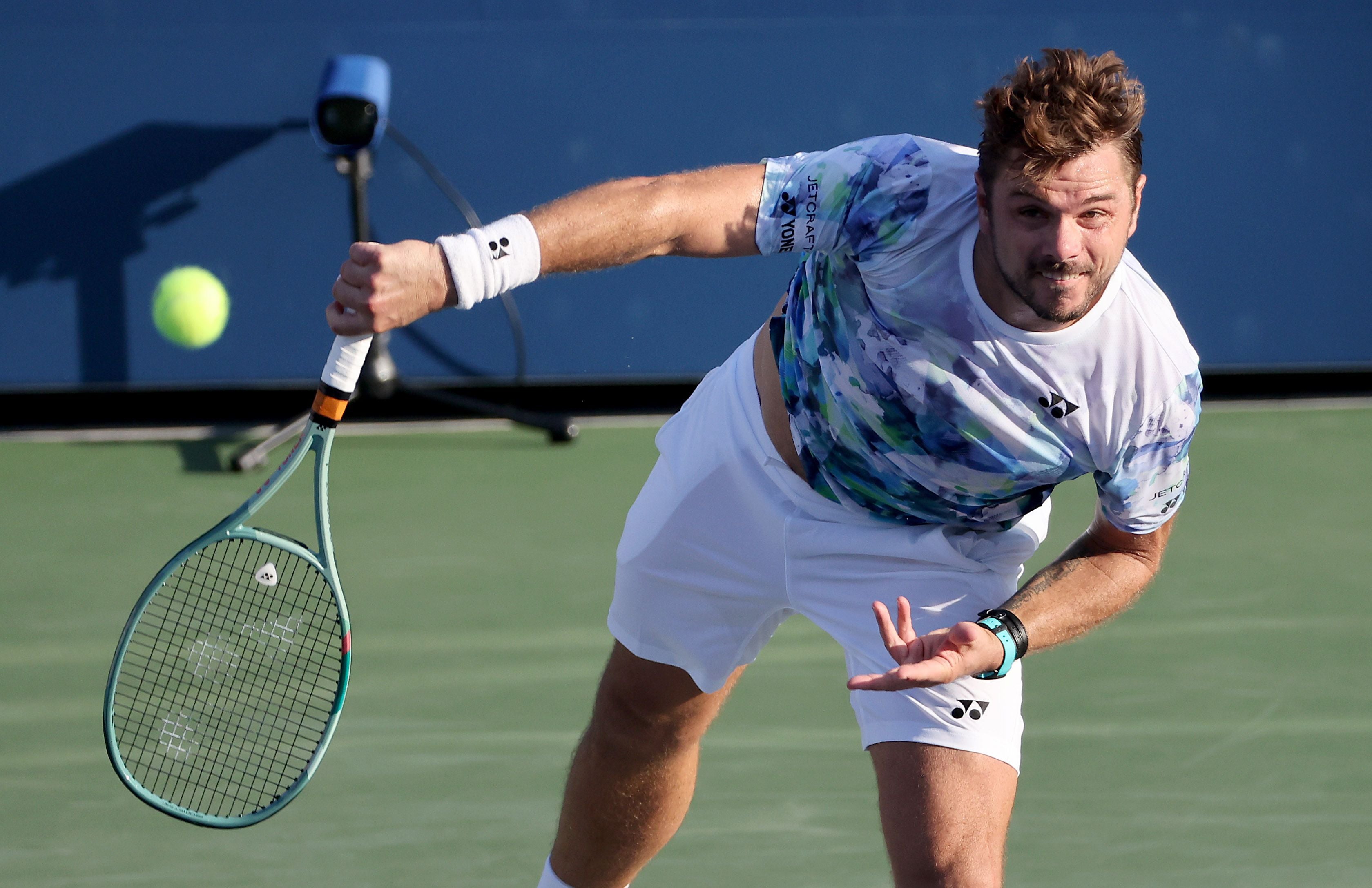 Tennisspieler Stan Wawrinka in weiß-blauem Trikot und weißer Hose setzt auf einem Hartplatz gebückt zu einer Vorhand an, während der gelbe Tennisball links im Bild durch die Luft fliegt.