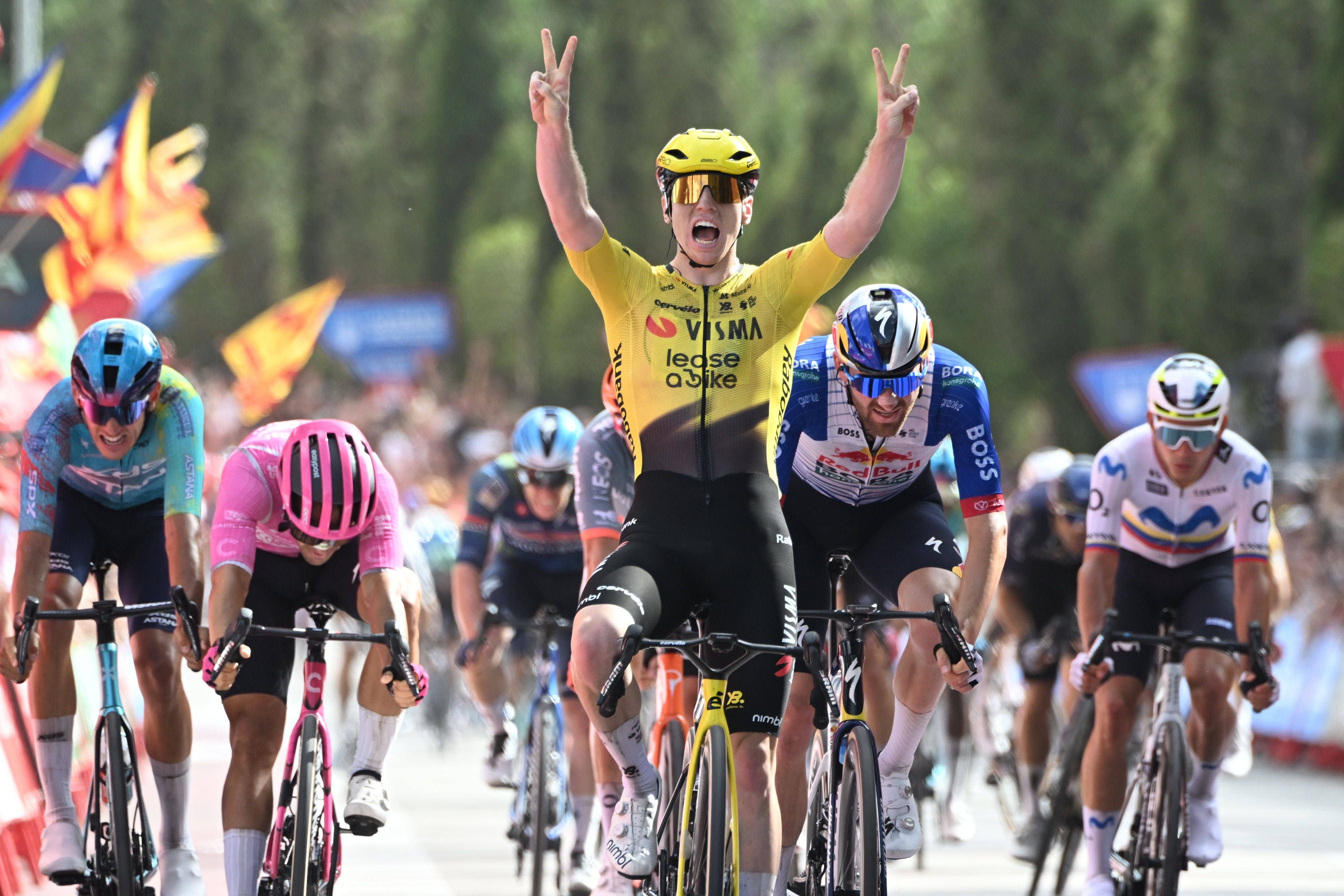 Matthew Brennan in gelbem Visma-Trikot fährt mit erhobenen Armen und Siegergeste über die Ziellinie, während hinter ihm weitere Sprinter auf ihren Rädern nachsetzen.