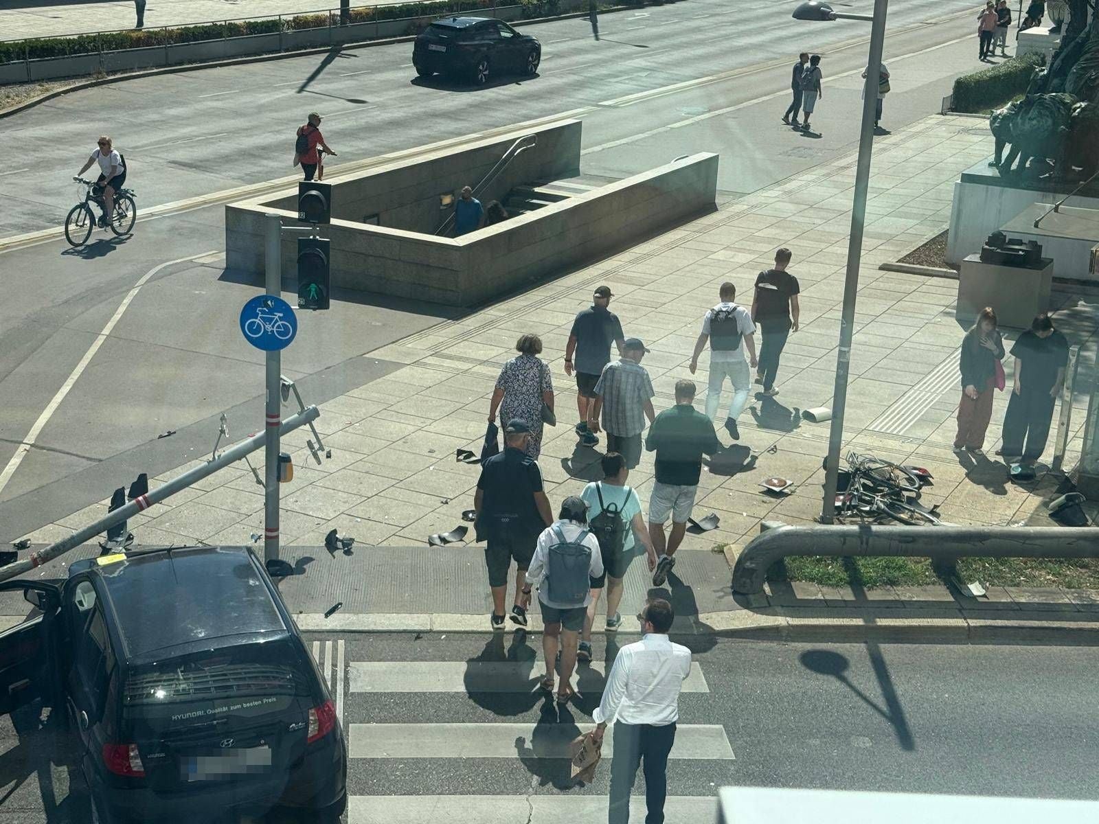 Menschen überqueren einen Zebrastreifen an einer Unfallstelle mit beschädigtem Auto und umgekipptem Mast.