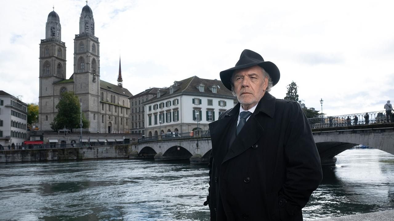 Älterer Mann im Mantel und Hut steht an einem Fluss vor dem Zürcher Grossmünster.