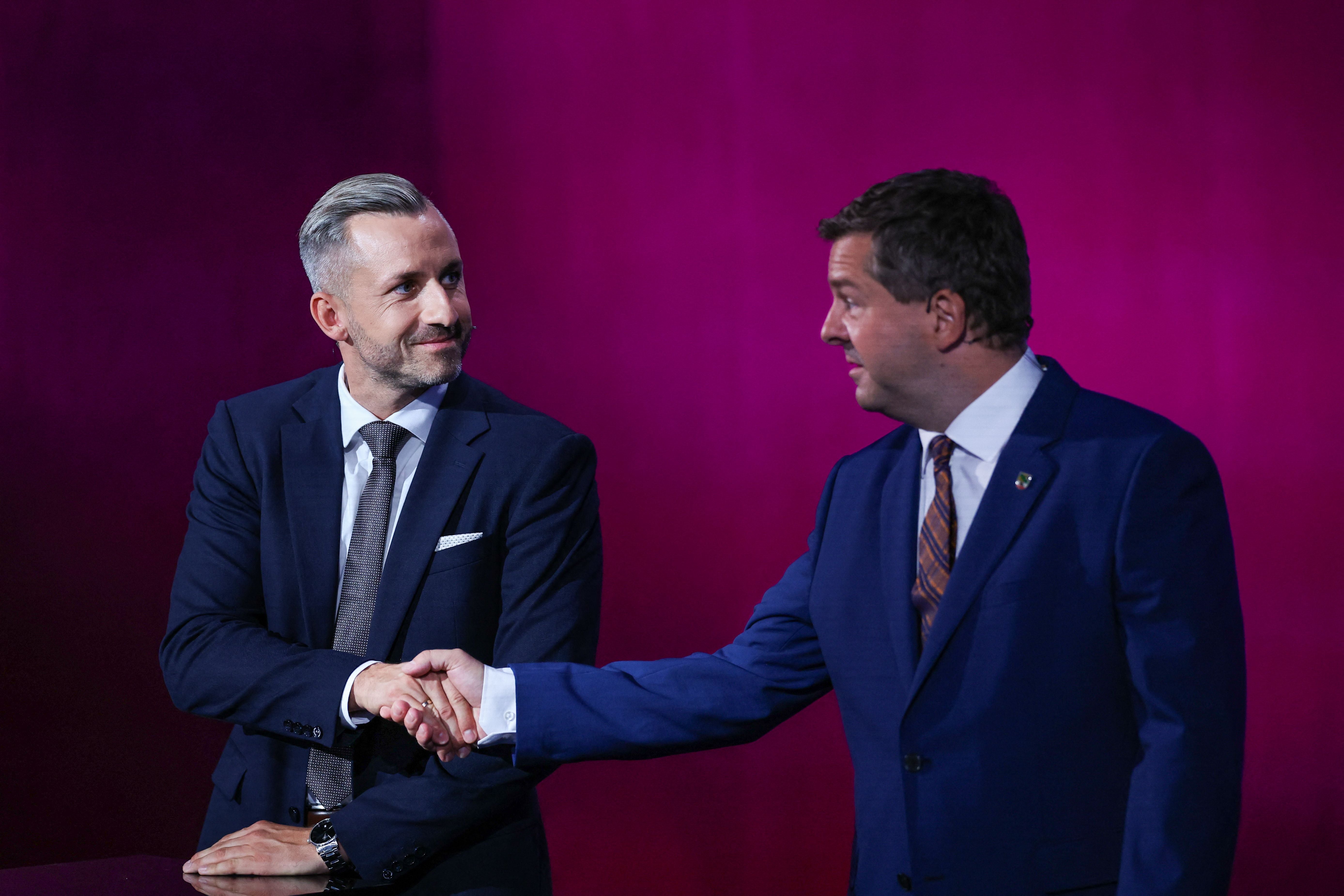 Zwei Männer im Anzug schütteln sich vor violettem Hintergrund die Hand und blicken sich an.