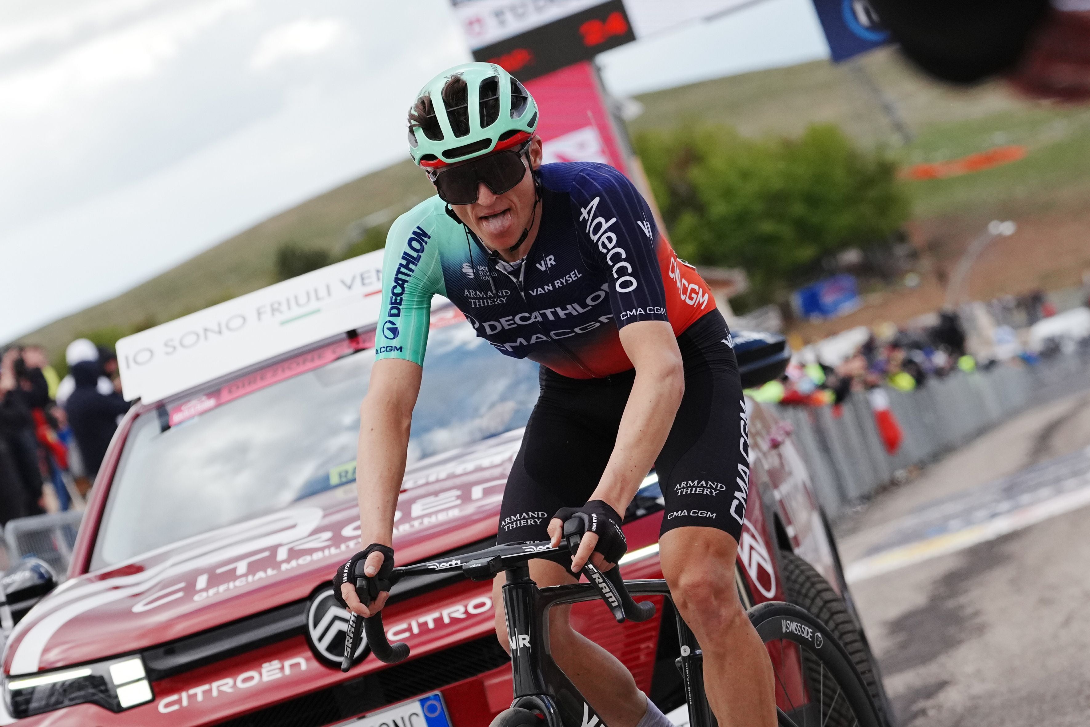 Felix Gall im dunklen Decathlon-Trikot mit hellblau-grünen Ärmeln fährt leicht gebeugt auf seinem Rennrad eine Steigung hinauf und streckt dabei die Zunge heraus. Im Hintergrund ist ein rotes Begleitfahrzeug zu sehen.