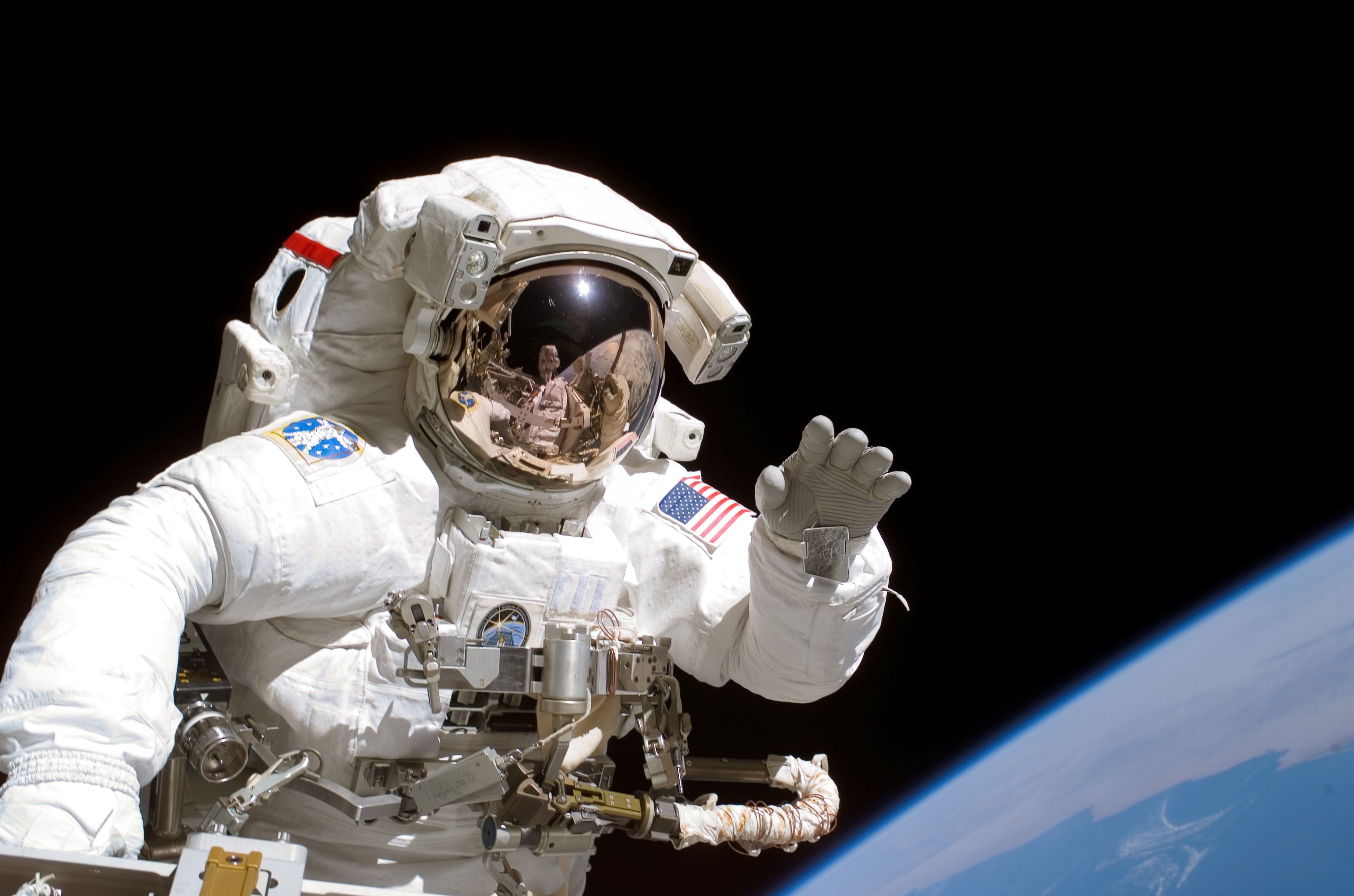 Ein Astronaut schwebt bei einem Weltraumspaziergang vor der Erde und winkt der Kamera.