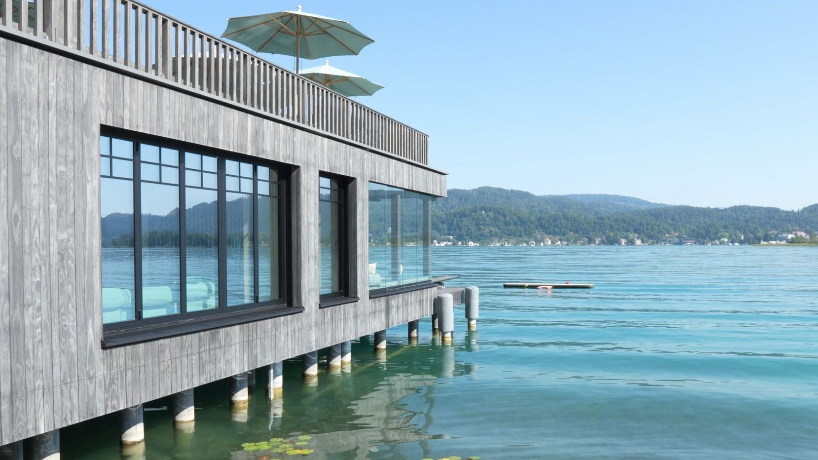 Modernes Gebäude mit Holzfassade steht auf Pfählen direkt am klaren Wasser eines Sees mit Bergkulisse.