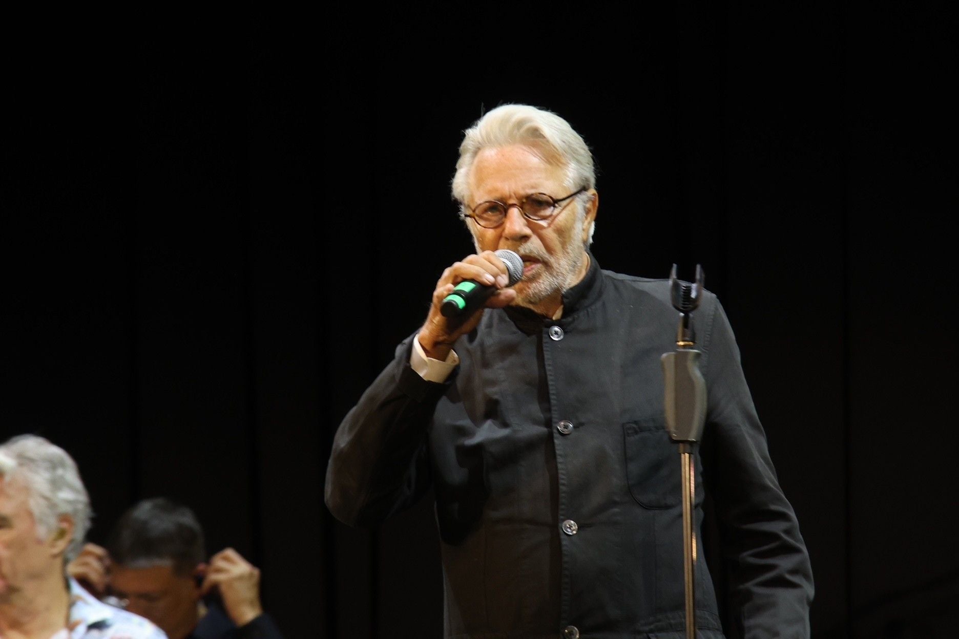 Ein älterer Mann mit Brille singt auf einer Bühne in ein Mikrofon.