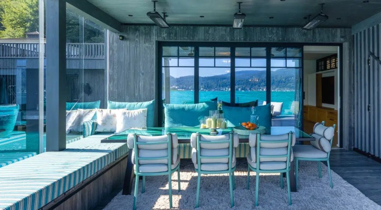 Modernes Esszimmer mit Glaswand, Seeblick, türkisfarbenen Stühlen und Bank mit gemusterten Kissen.