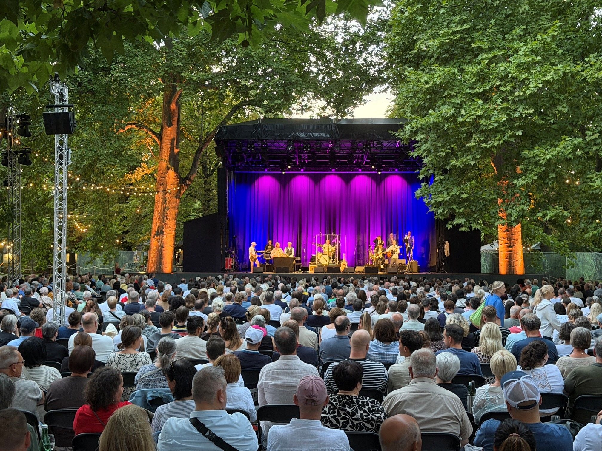 Open-Air-Konzert mit großer Menschenmenge vor einer beleuchteten Bühne im Park bei Abenddämmerung.