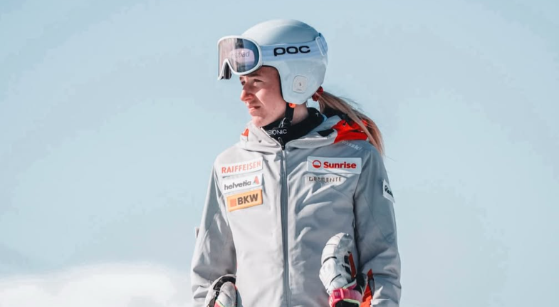 Skifahrerin mit Helm und Skibrille steht draußen im Schnee und blickt zur Seite.