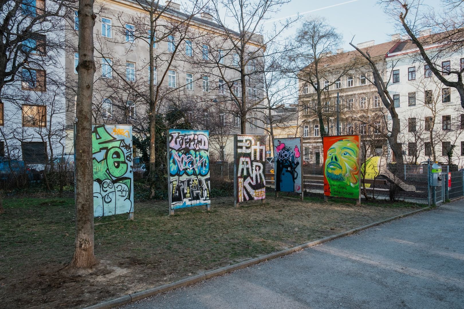 Mehrere bemalte Tafeln mit Graffiti und Porträt im H.C.-Artmann-Park vor Wohnhäusern.