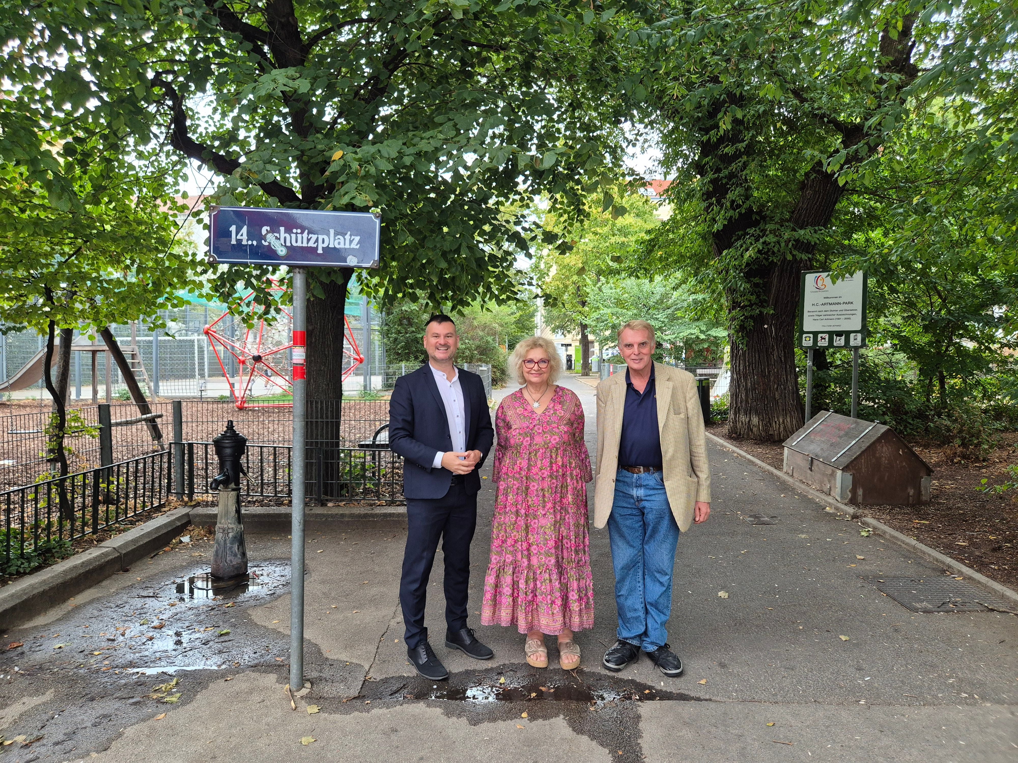 Drei Personen stehen im H.-C.-Artmann-Park neben einem Schild mit der Aufschrift „14. Schützplatz“.