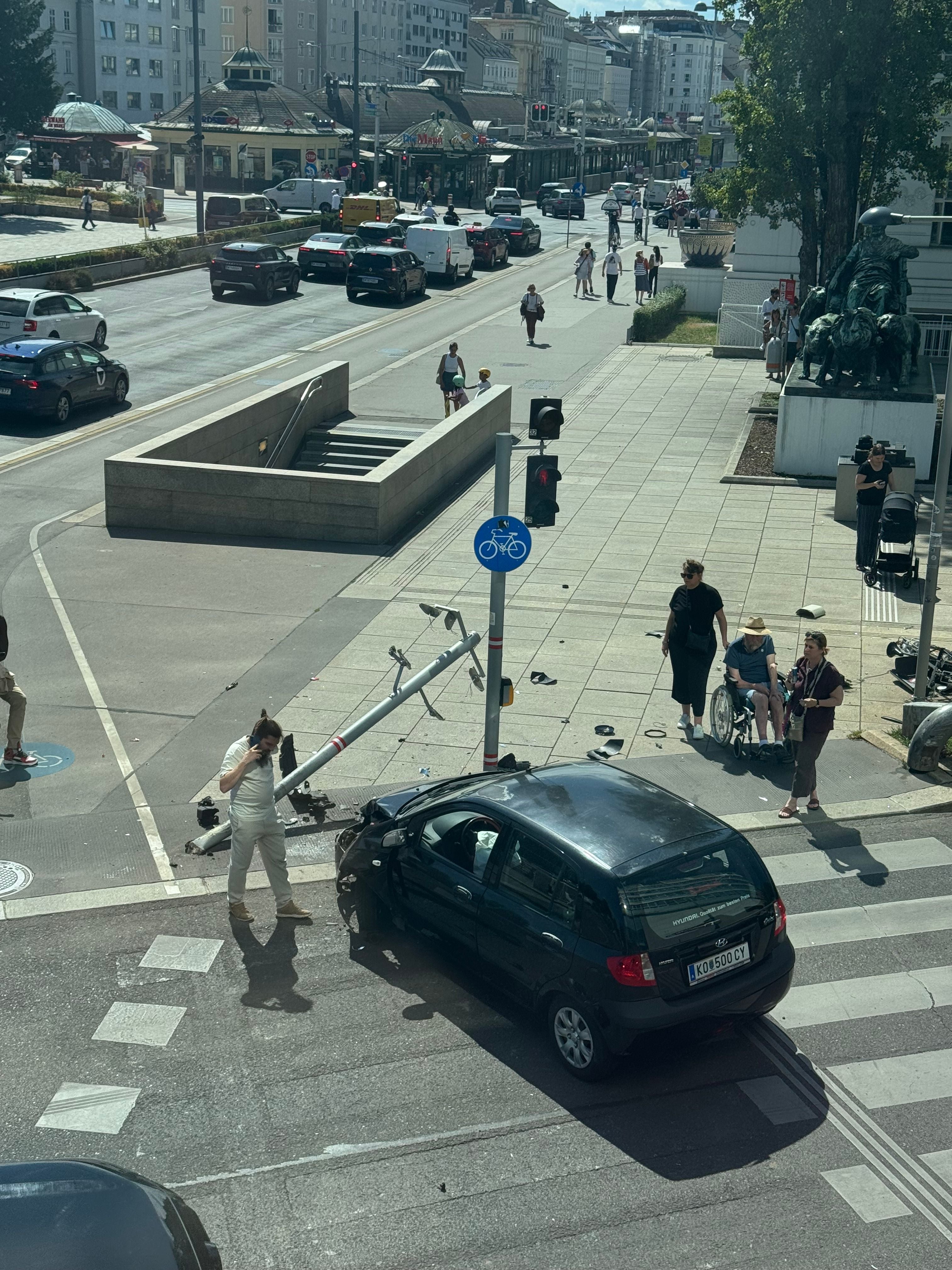 Schwarzes Auto nach Unfall mit umgefahrener Ampel an einer Kreuzung, Personen stehen daneben.