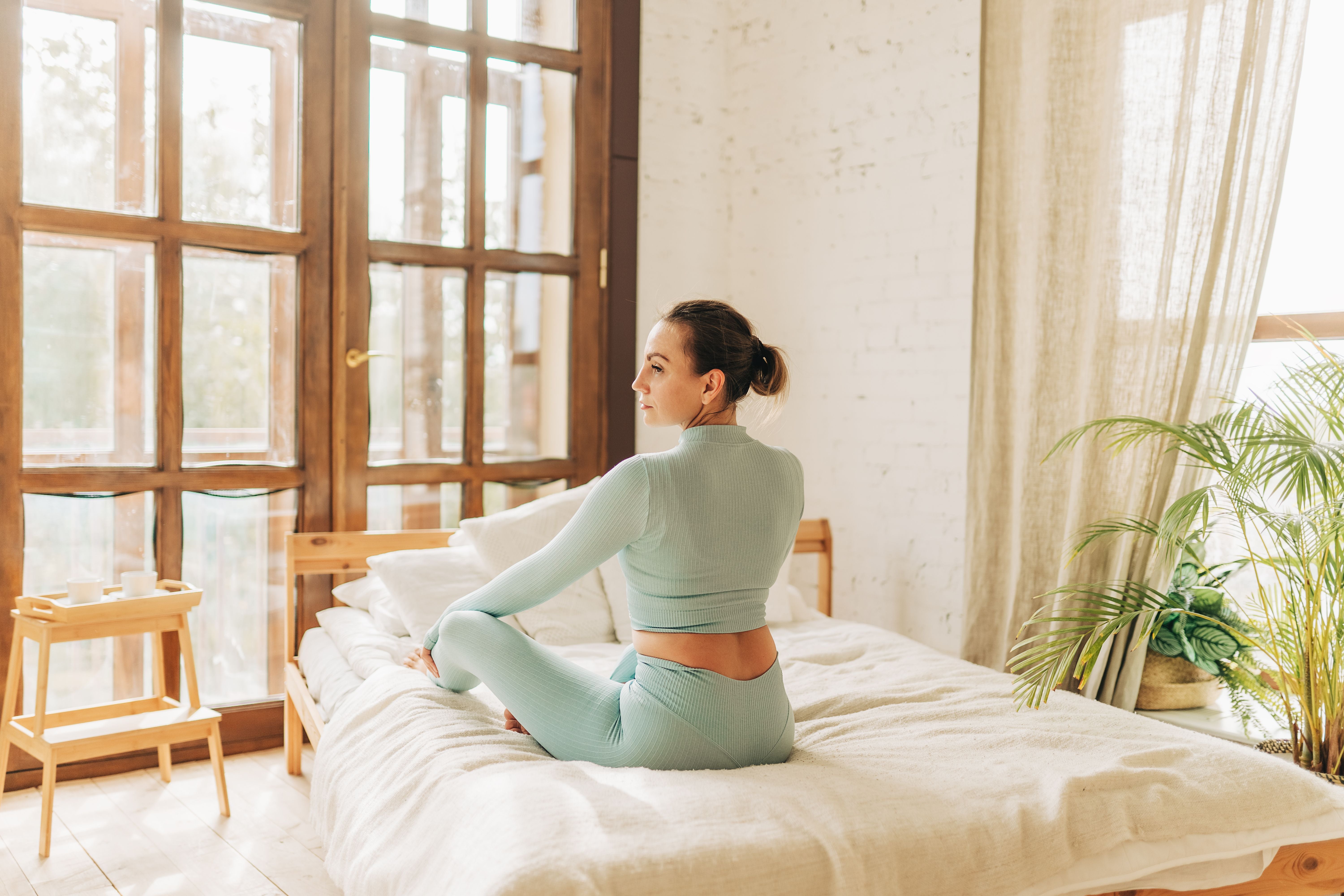 Frau in Sportkleidung macht Yoga auf dem Bett in einem hellen Schlafzimmer.