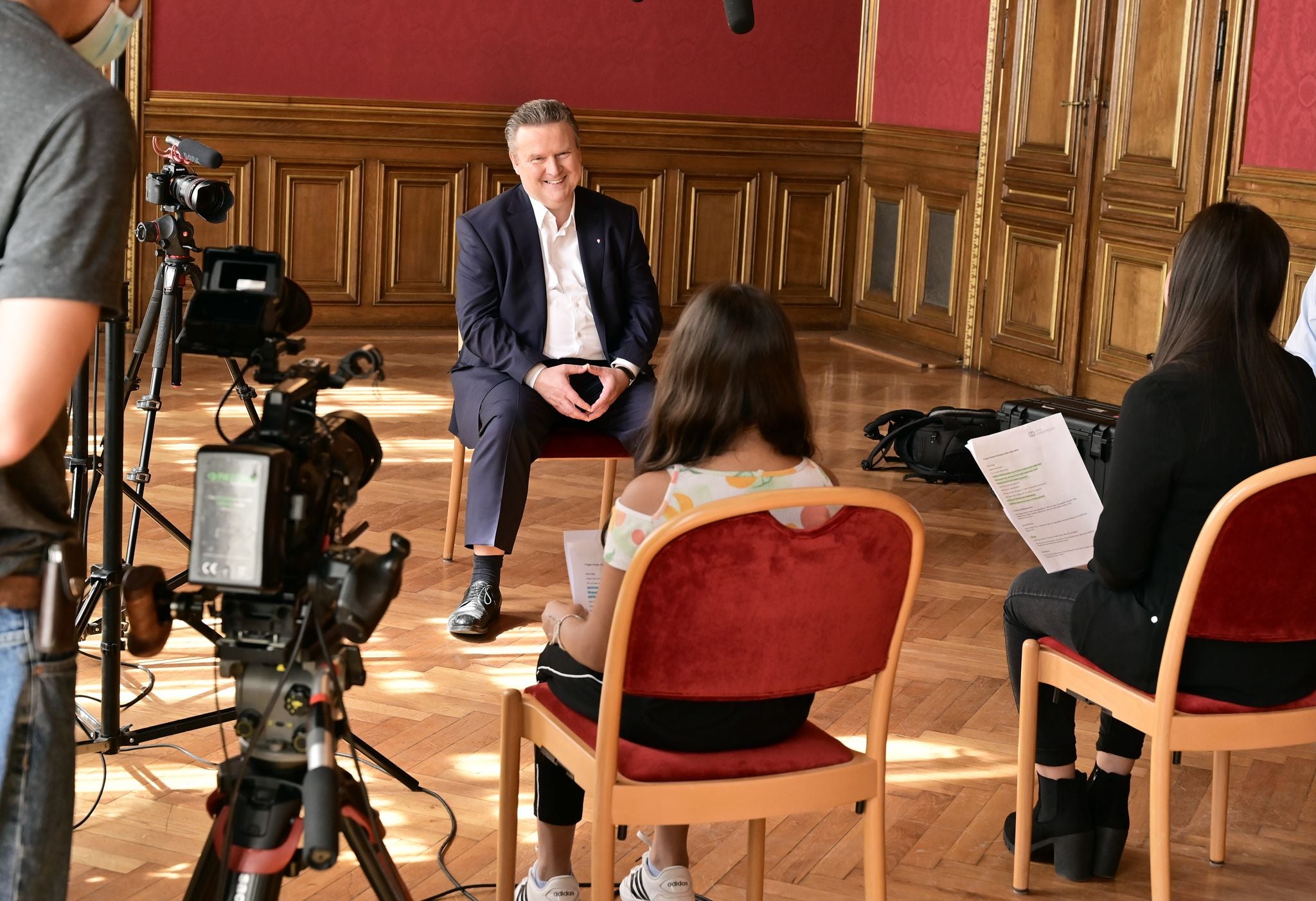 Interview mit einem Mann im Anzug, gefilmt von Kameras, zwei Frauen sitzen ihm gegenüber.
