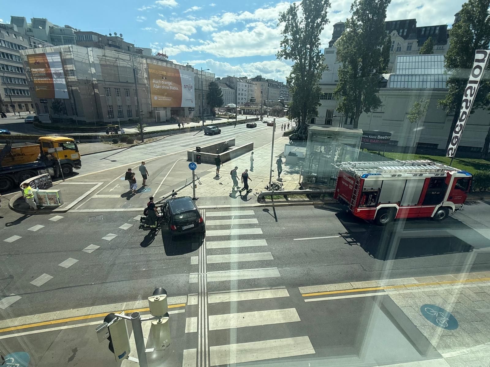 Feuerwehrwagen und Auto an Kreuzung, mehrere Menschen, sonniger Tag, Blick aus dem Fenster.