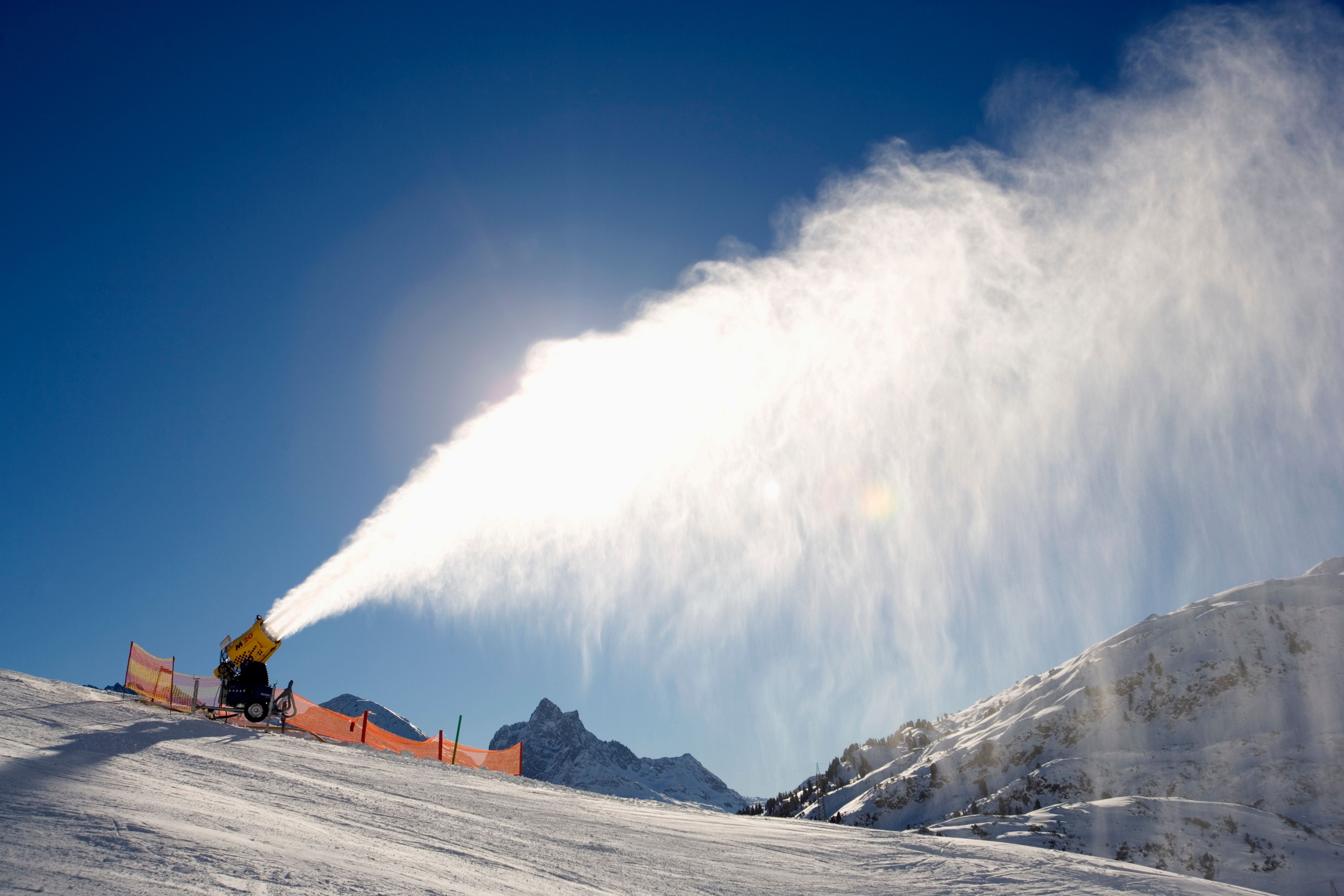 Eine Schneekanone produziert künstlichen Schnee auf einer Skipiste bei sonnigem Wetter.