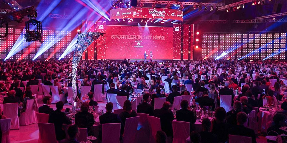 Großer Festsaal mit vielen Gästen bei einer Gala, Bühne mit roter Beleuchtung im Hintergrund.