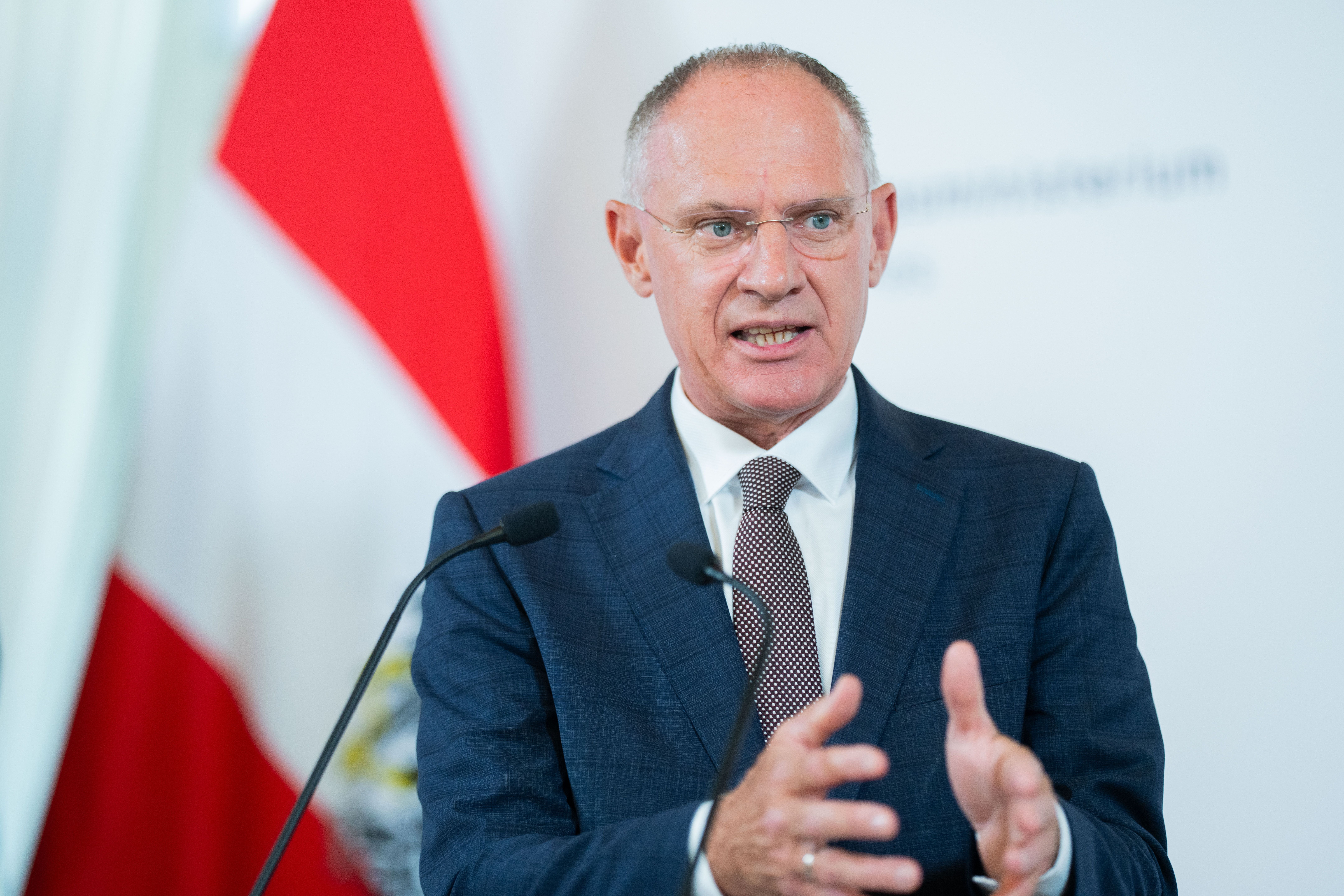 Ein Mann im Anzug spricht bei einer Pressekonferenz mit Mikrofonen vor einer Flagge.