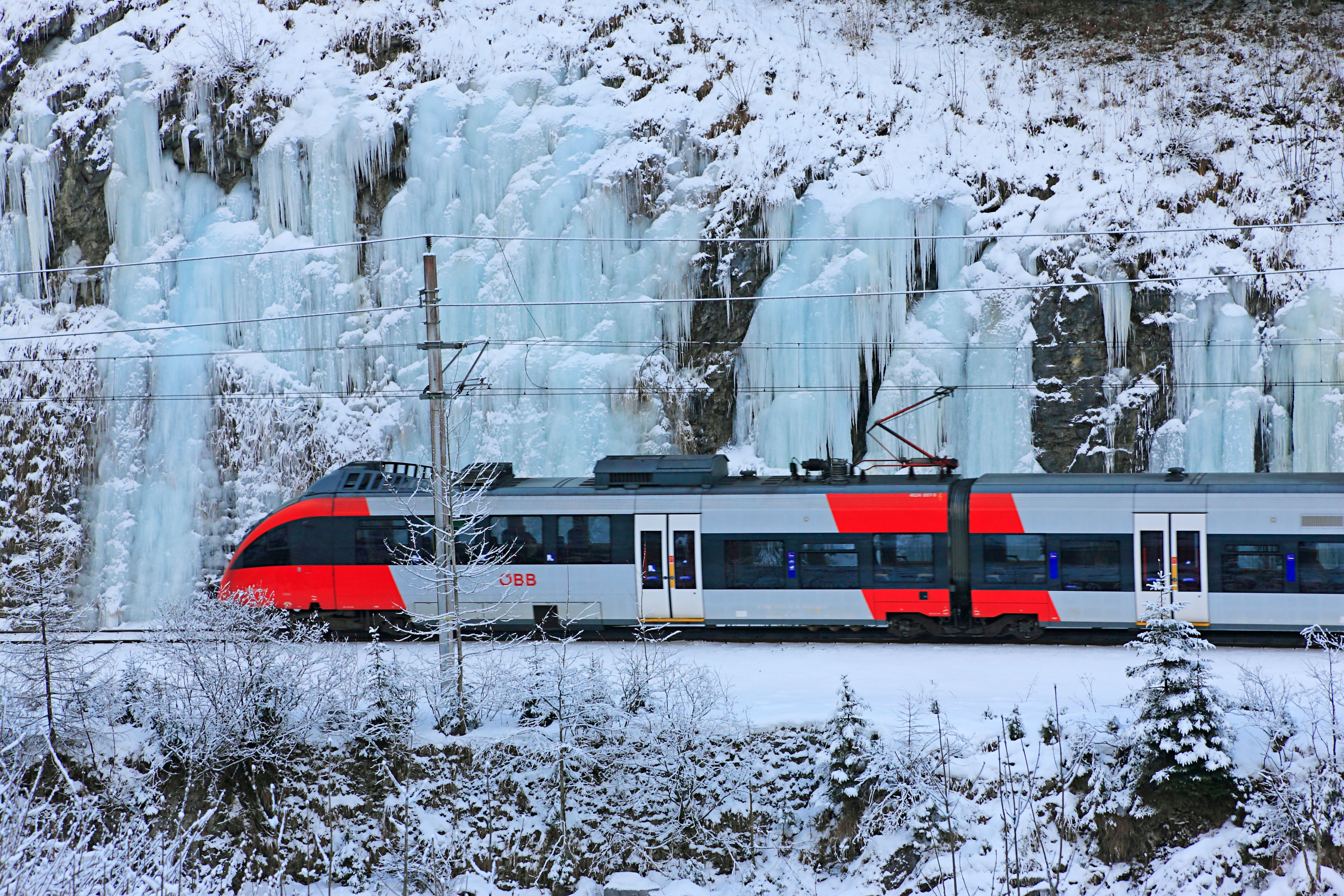 Ein moderner Zug fährt vor einer eisbedeckten Felswand durch eine verschneite Winterlandschaft.