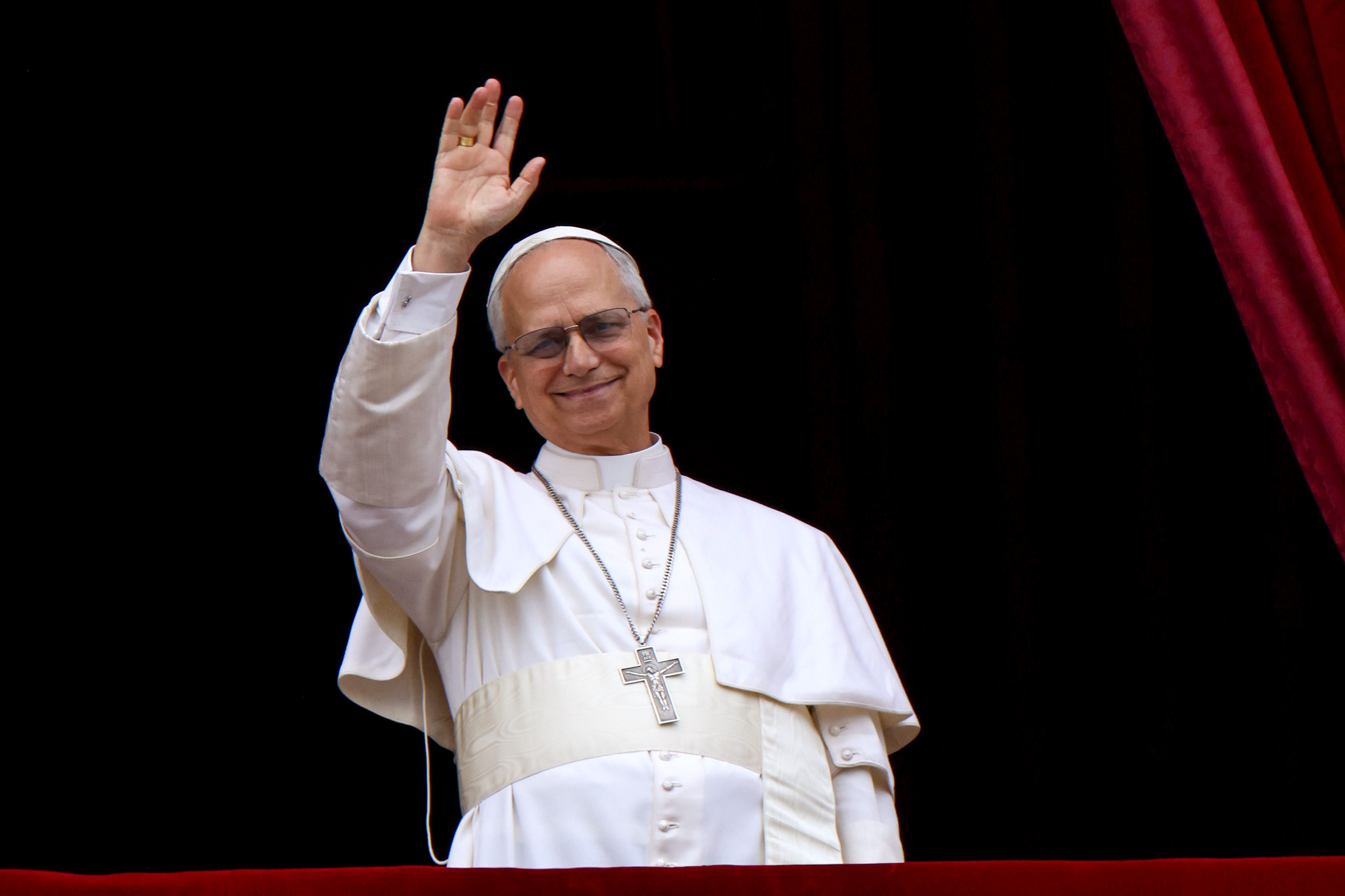 Ein Mann im weißen Papstgewand winkt lächelnd von einem Balkon mit rotem Vorhang.