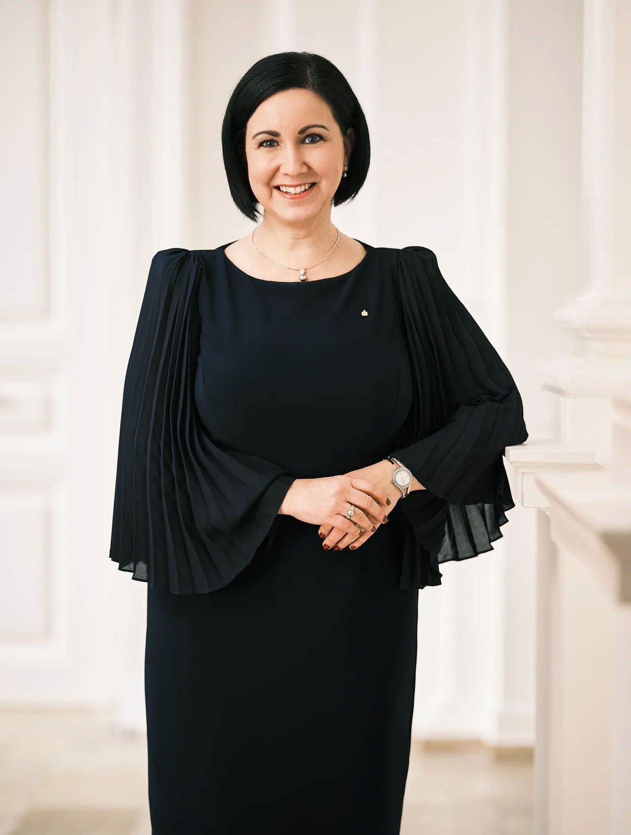 Frau mit schwarzem Kleid und dunklen Haaren steht lächelnd in elegantem, hellen Innenraum.