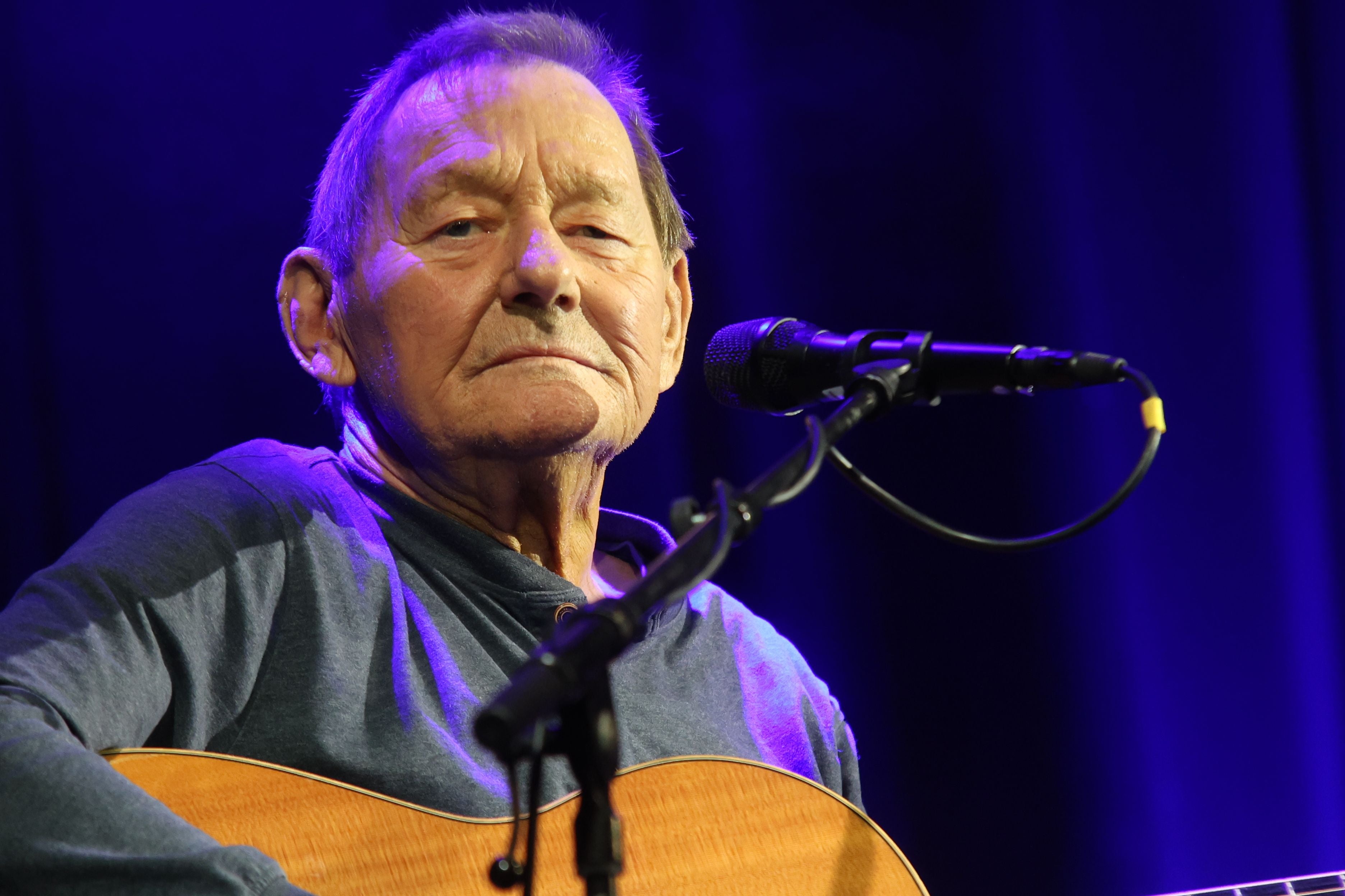 Älterer Mann spielt Gitarre und singt vor Mikrofon auf einer Bühne mit blauem Licht.