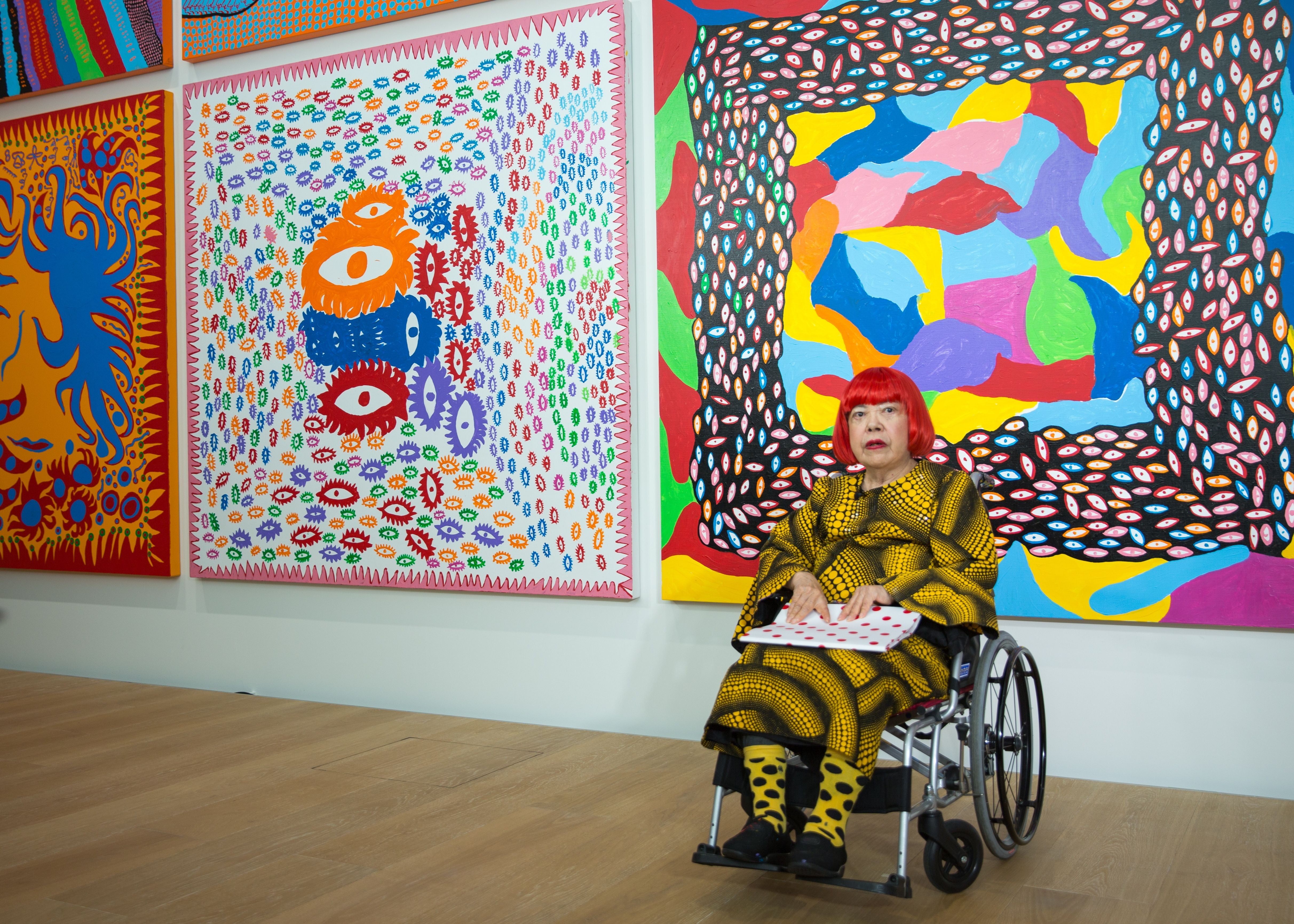 Eine Frau mit rotem Bob sitzt im Rollstuhl vor bunten, abstrakten Gemälden in einer Galerie.