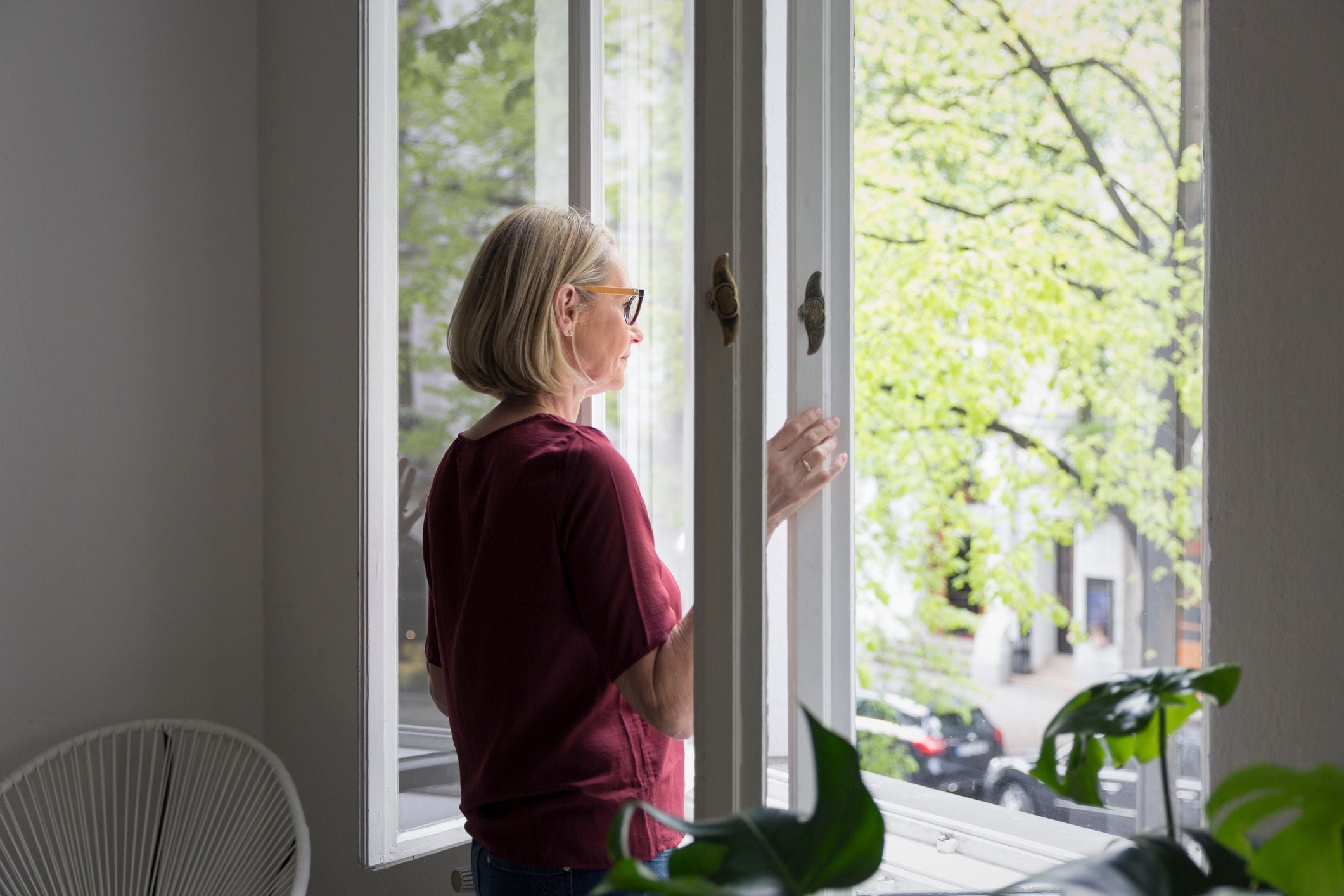 Frau mit Brille schaut nachdenklich aus einem geöffneten Fenster ins Grüne.