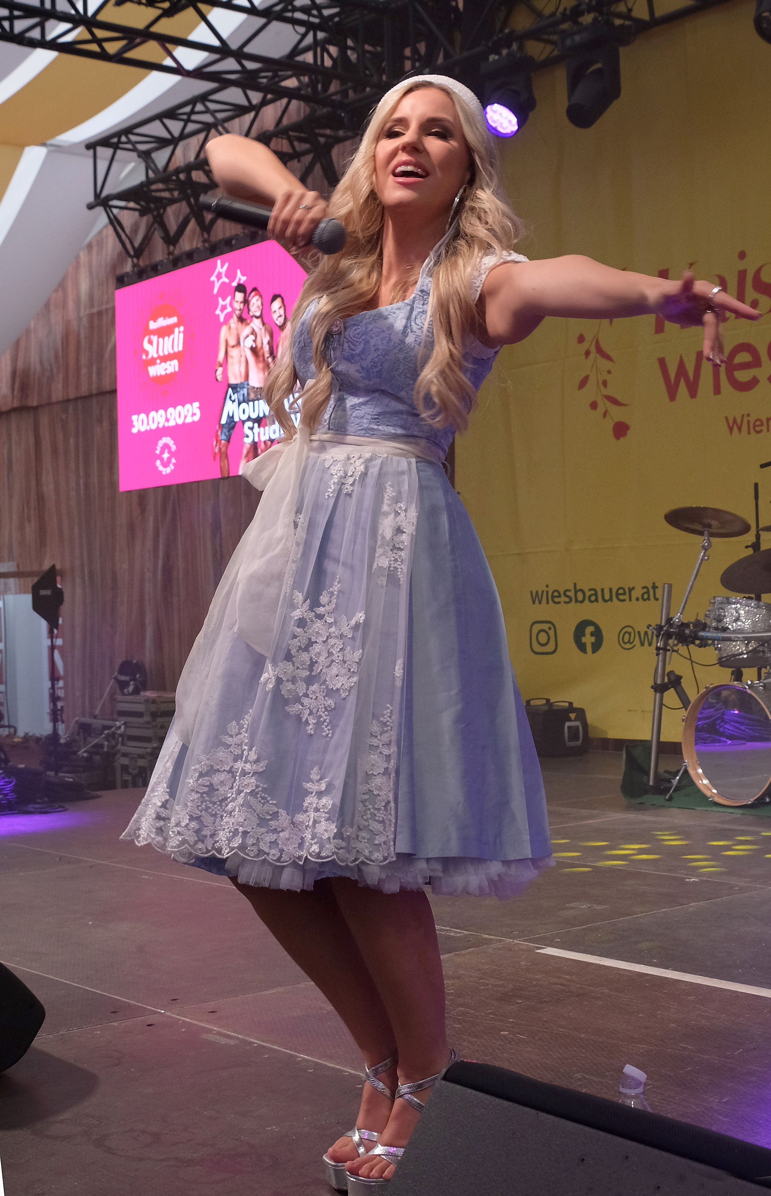 Eine Frau im blauen Dirndl singt auf einer Bühne mit Mikrofon und breitet die Arme aus.