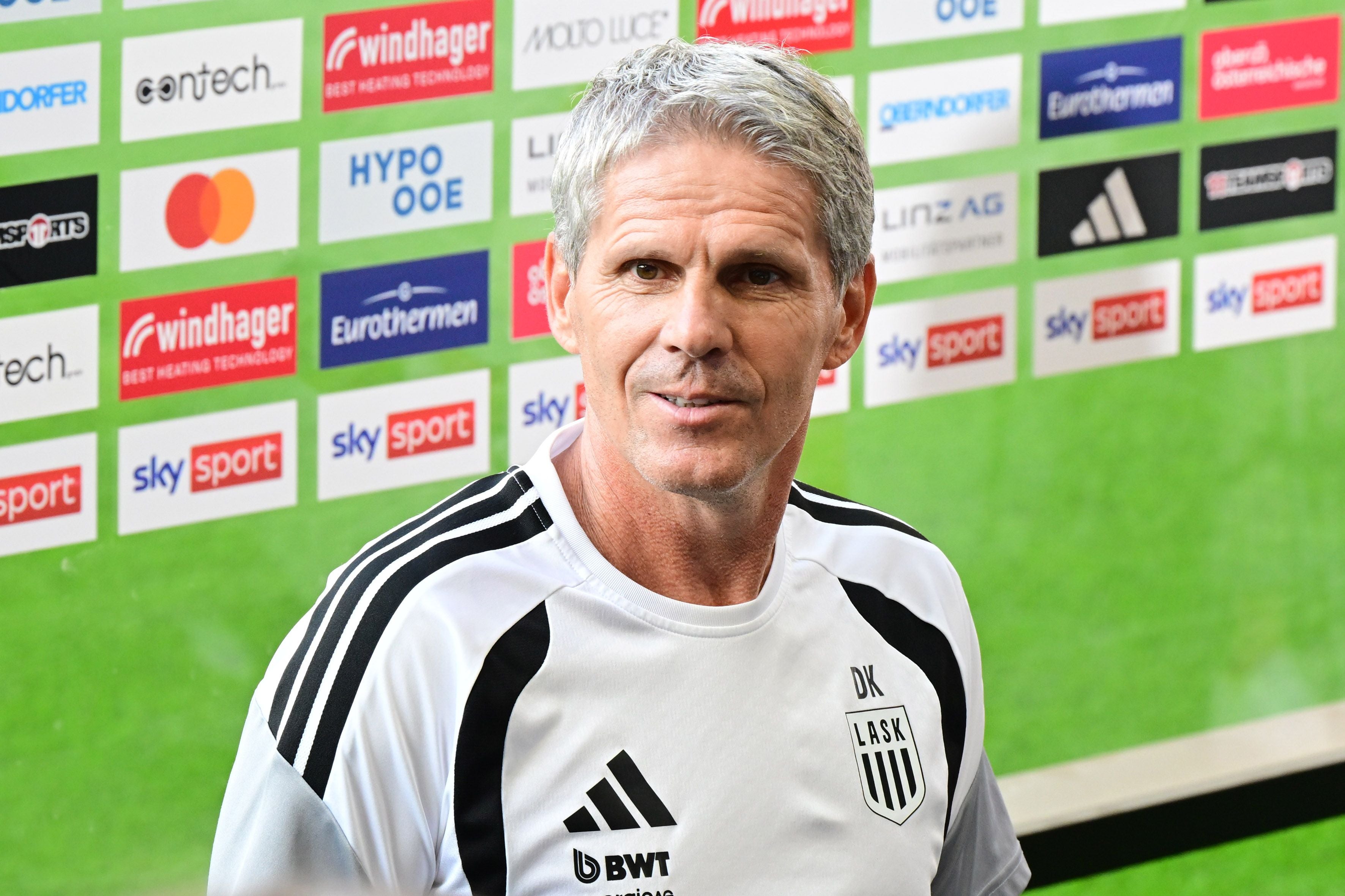 Ein Mann im LASK-Trainingsanzug steht vor einer Sponsorenwand im Fußballstadion.