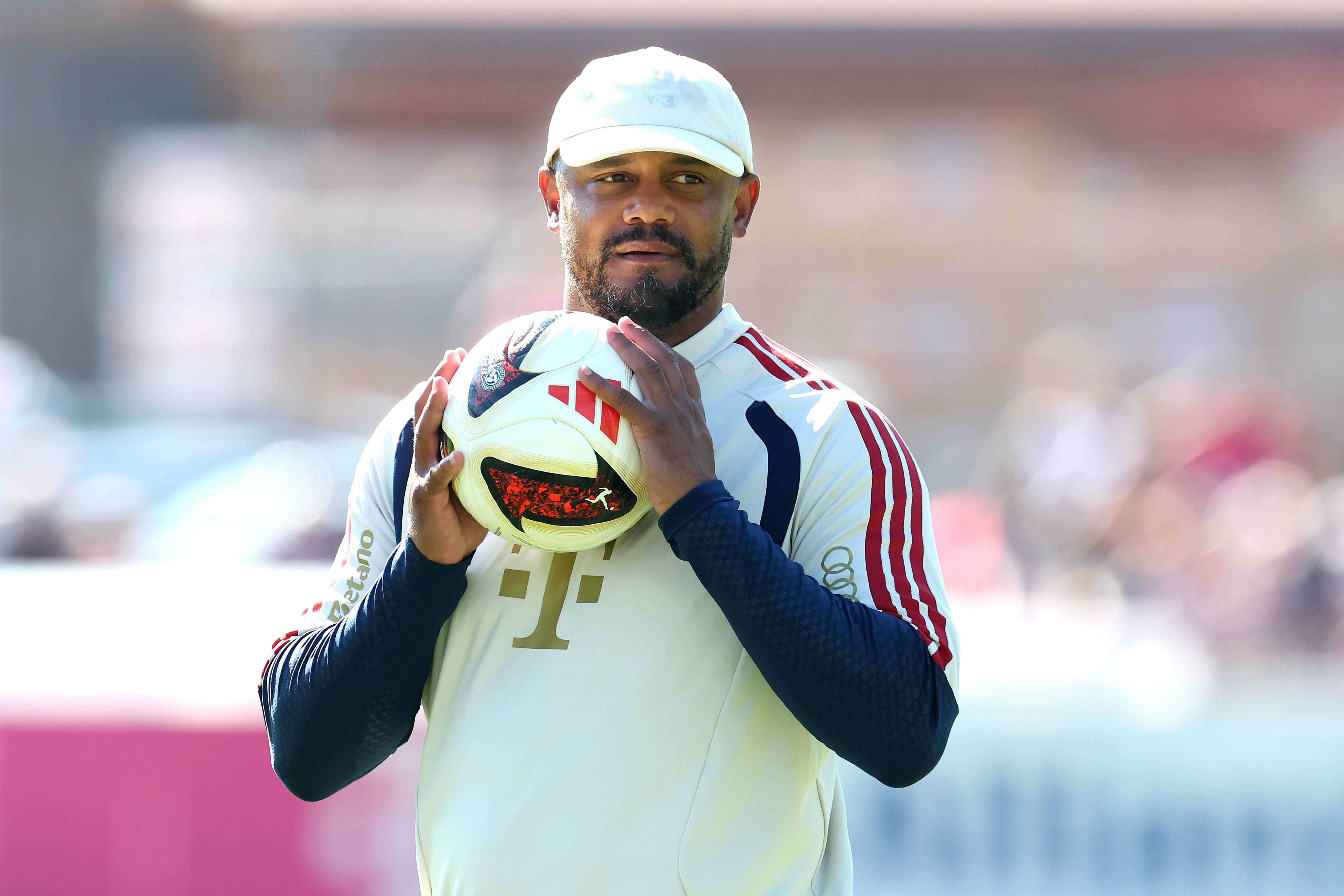 Ein Mann im Trainingsanzug hält einen Fußball und trägt eine weiße Kappe im Freien.