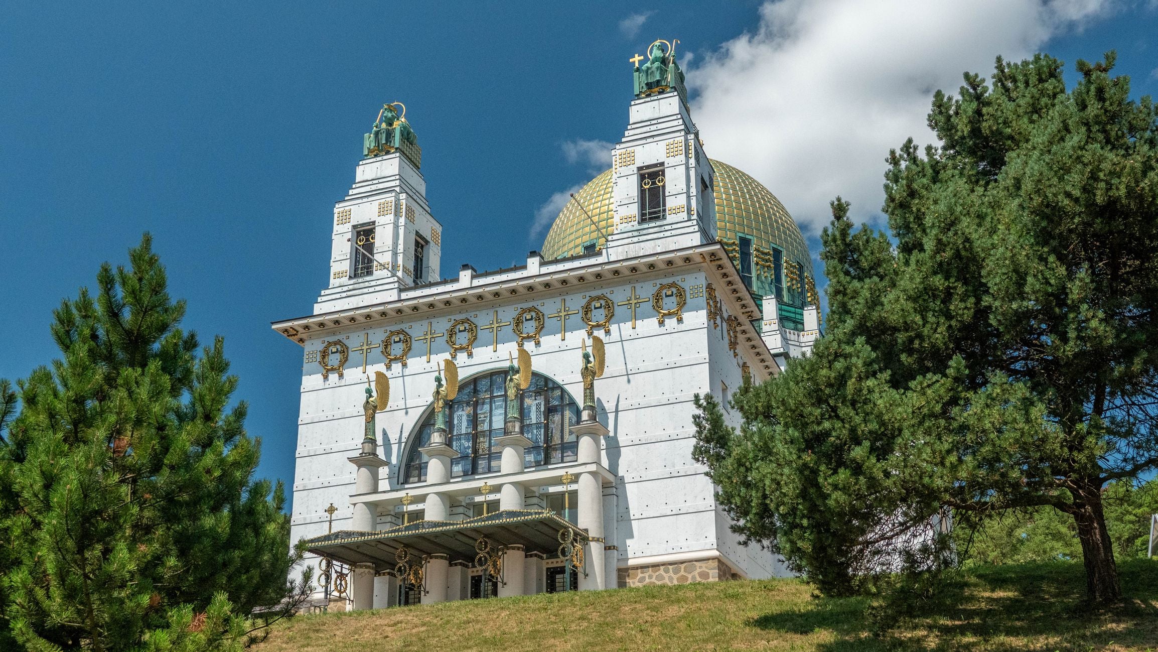 Die Kirche am Steinhof in Wien mit goldener Kuppel, Fassade im Jugendstil, umgeben von Bäumen.