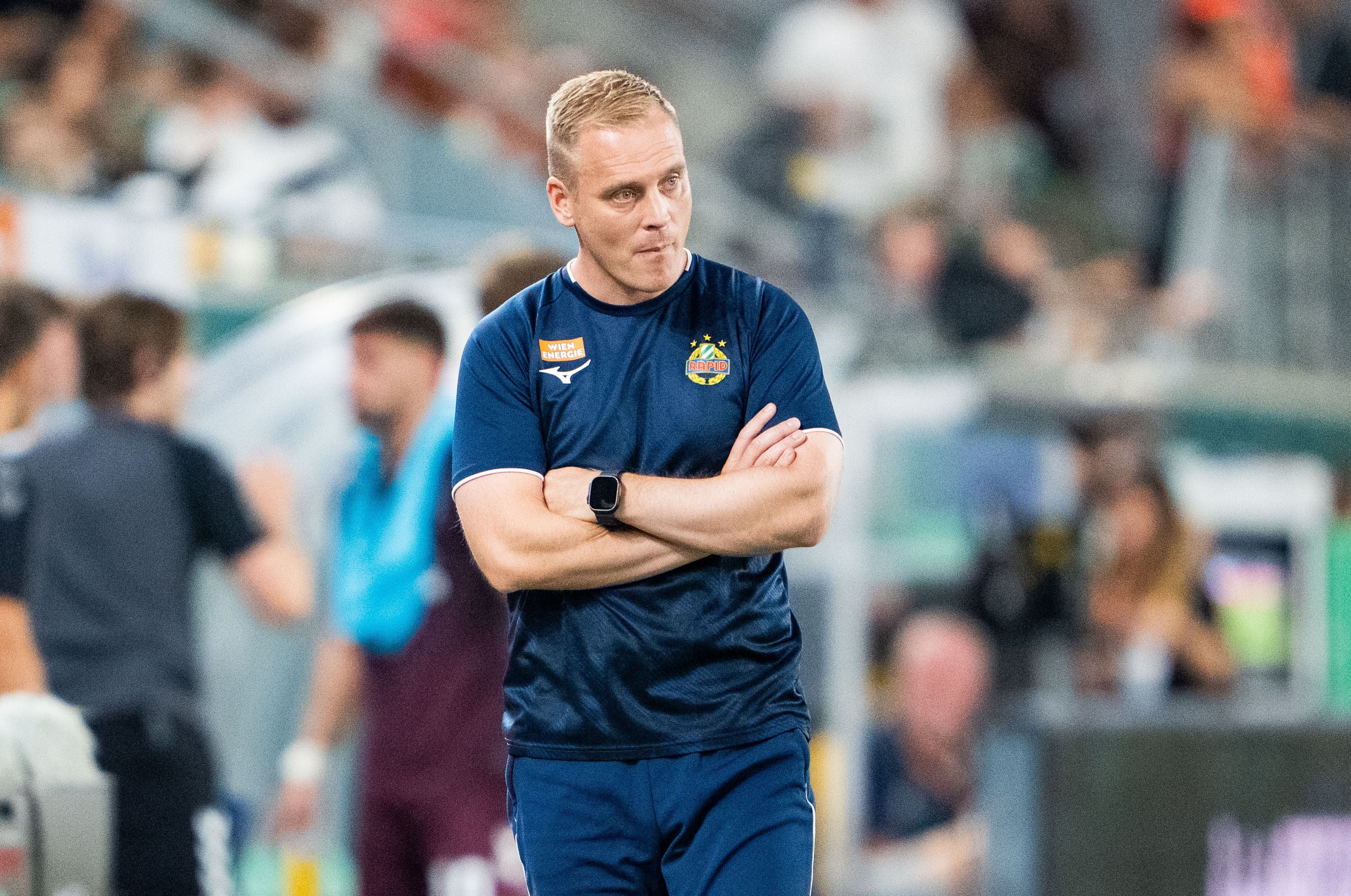 Ein Fußballtrainer steht mit verschränkten Armen am Spielfeldrand während eines Spiels.