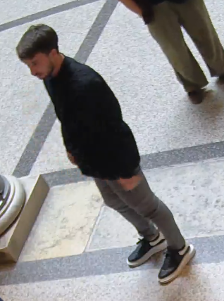 Junger Mann mit dunklem Pullover und grauer Hose steht auf gemustertem Boden.