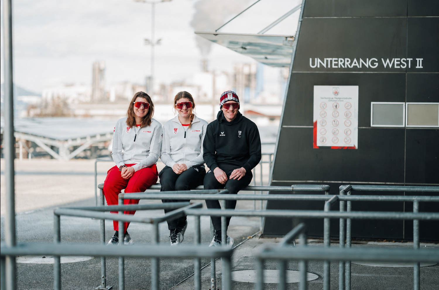 Drei Personen mit Sportkleidung und Sonnenbrillen sitzen vor einem Gebäude mit der Aufschrift "UNTERRANG WEST II".