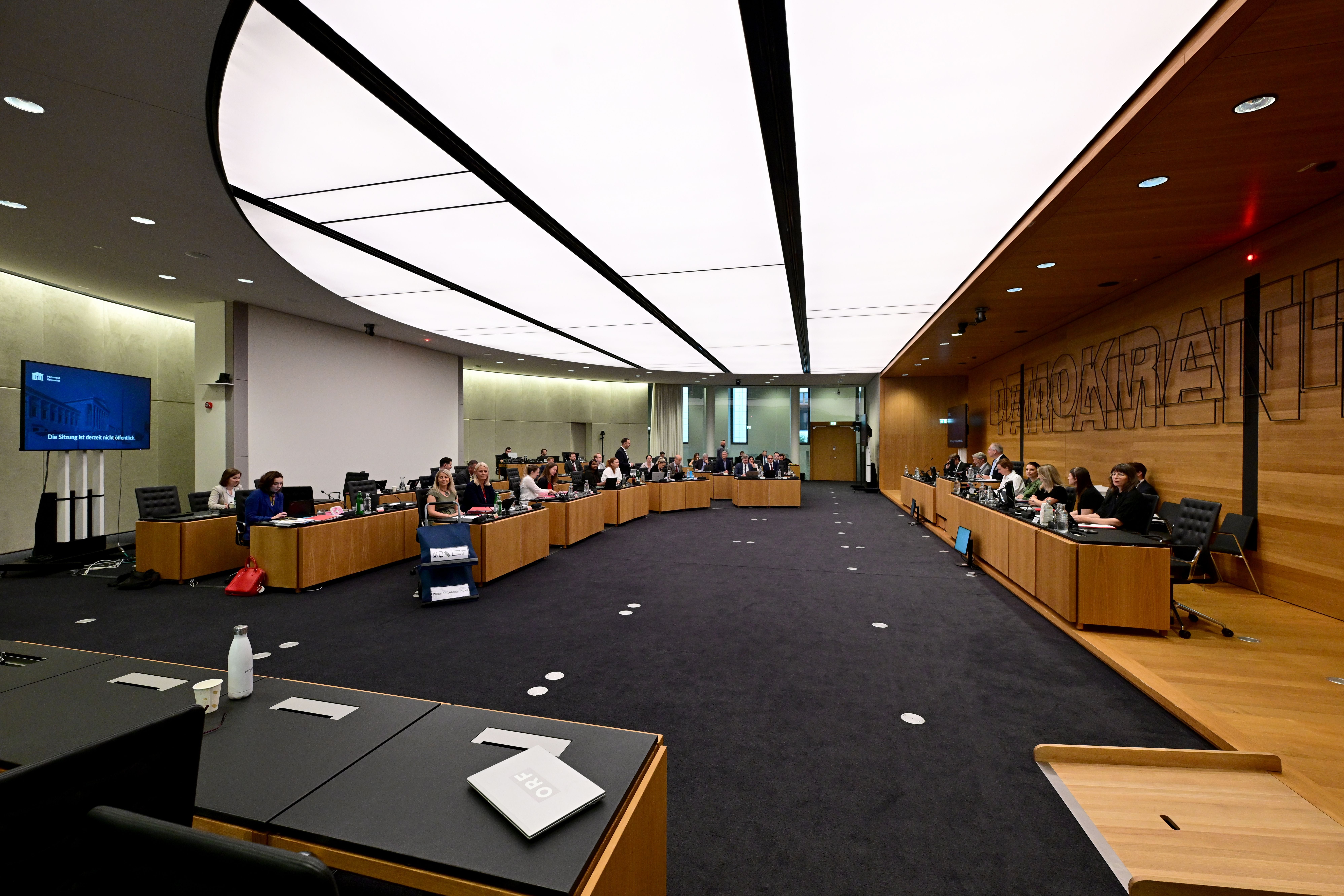 Großer Sitzungssaal im Parlament Wien mit mehreren Personen an langen Tischen vor einer Ausschusssitzung.