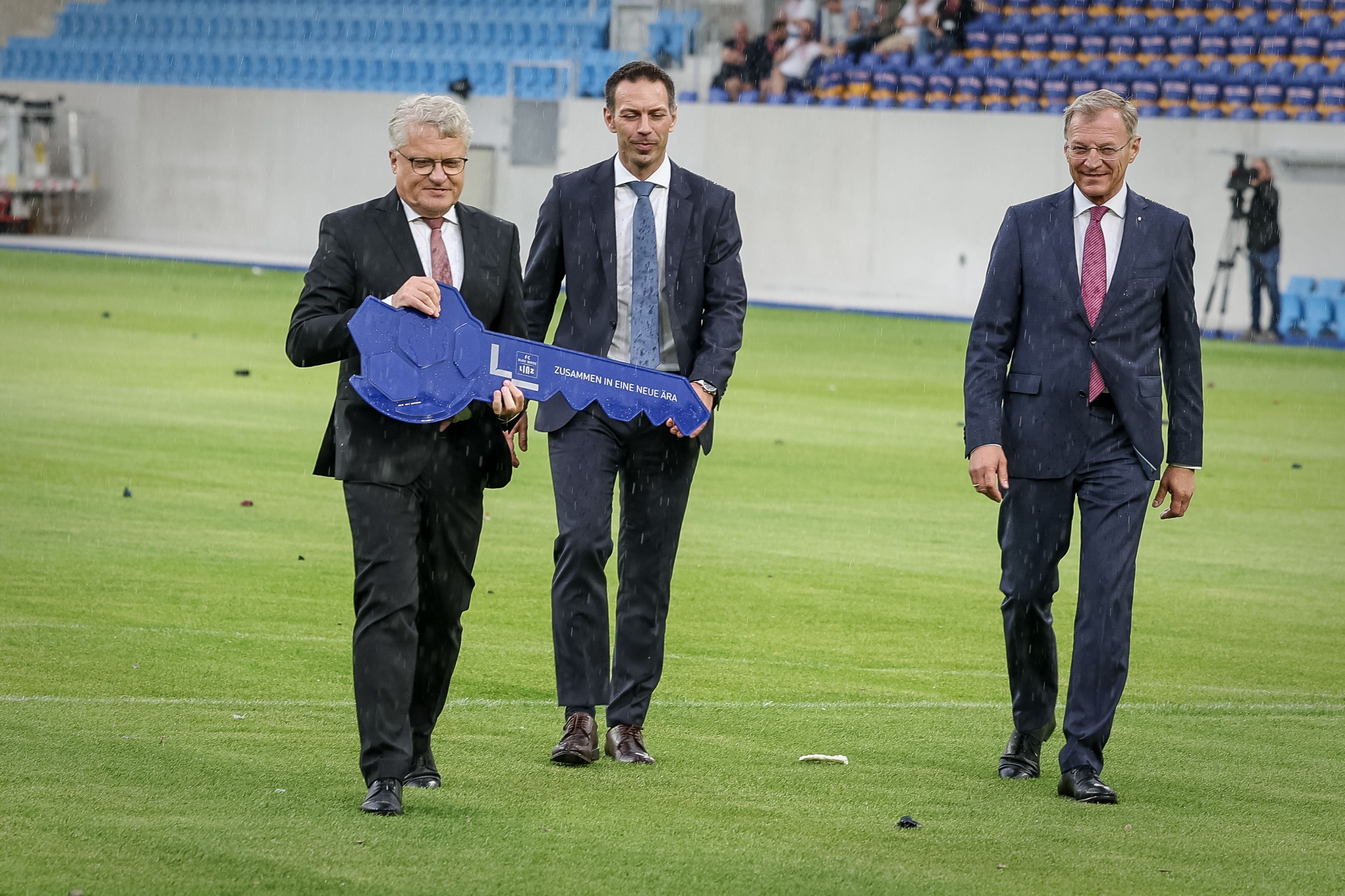 Drei Männer im Anzug gehen auf einem Fußballfeld, einer hält einen großen symbolischen Schlüssel.