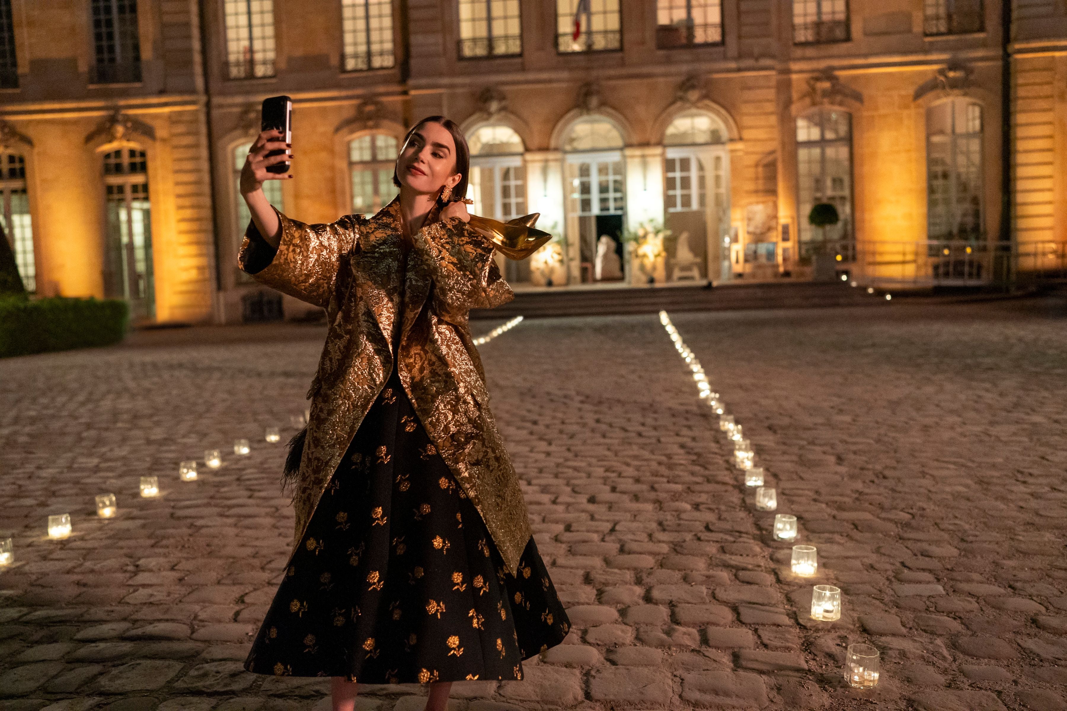 Frau in goldener Jacke macht ein Selfie mit goldfarbenem Schuh vor beleuchtetem Gebäude.