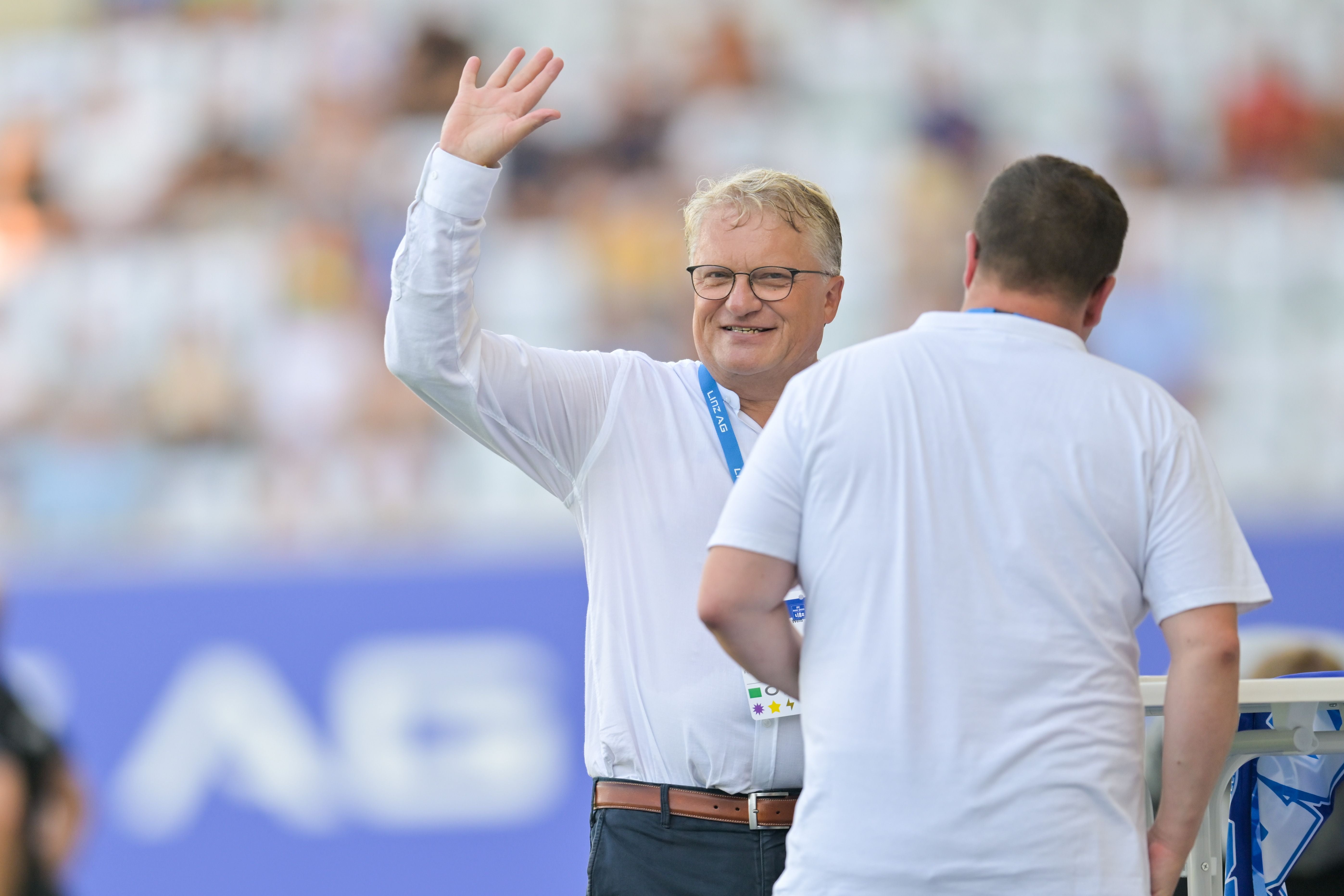 Ein Mann mit Brille winkt lächelnd in einem Stadion, eine weitere Person steht daneben.