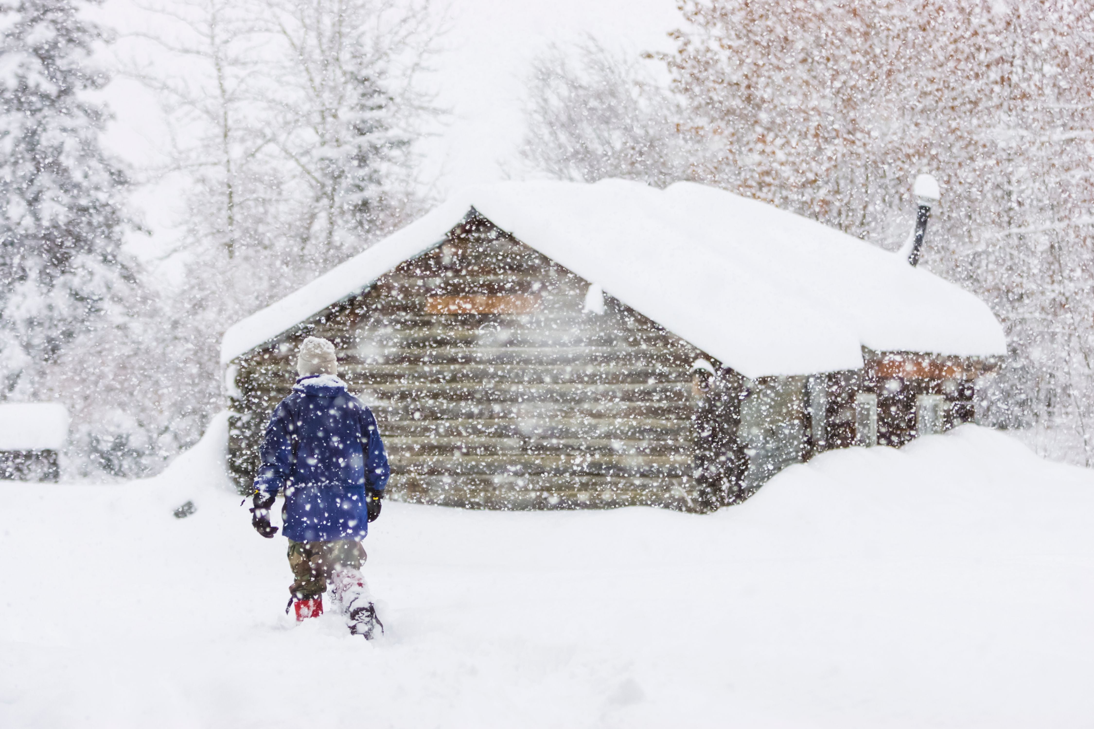 Person geht durch tiefen Schnee zu einer Blockhütte, es schneit stark.