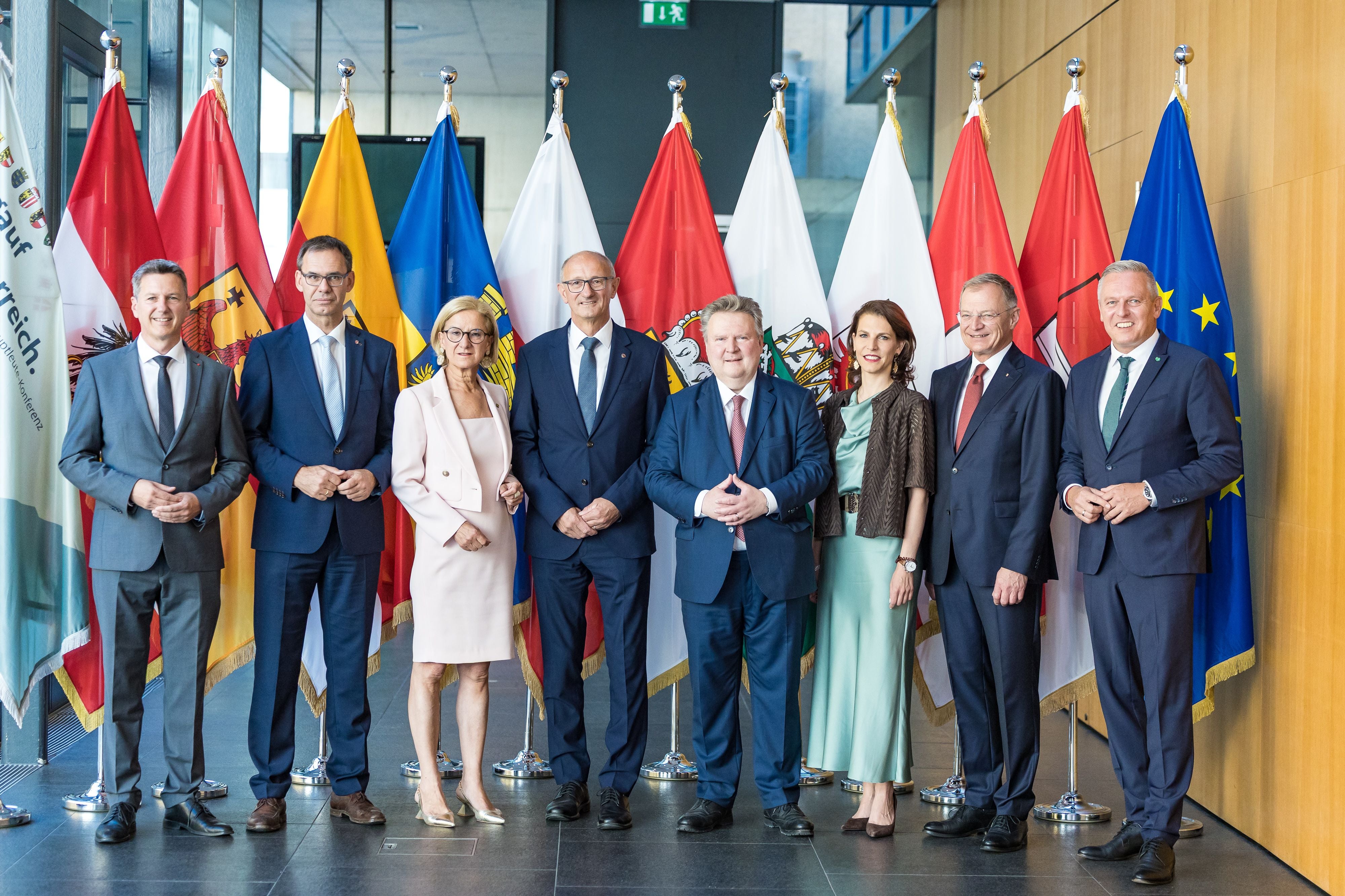 Acht Personen in Businesskleidung stehen vor Landes- und EU-Flaggen in einem modernen Gebäude.