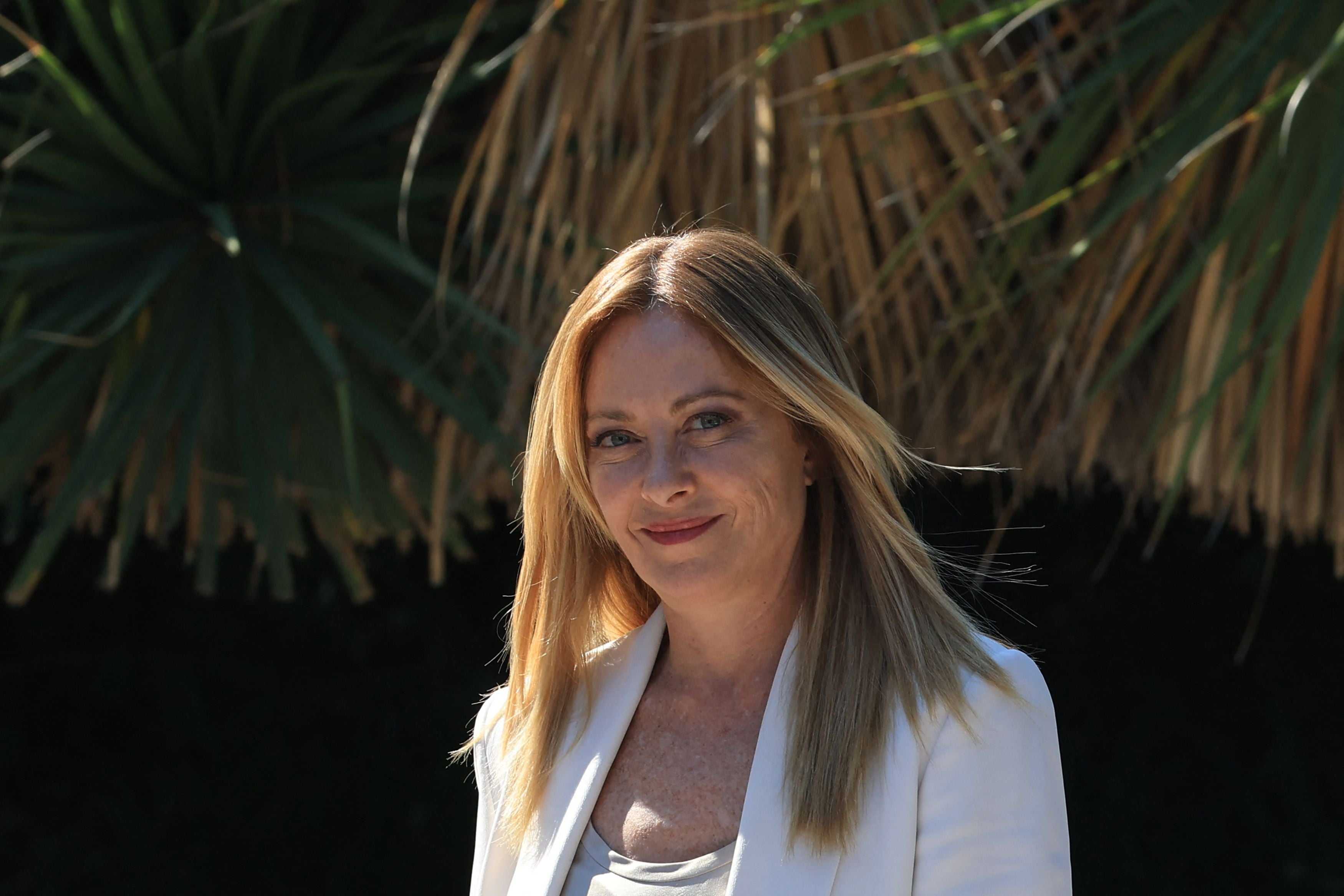 Frau mit langen blonden Haaren trägt weißen Blazer, im Hintergrund befindet sich grüne Vegetation.