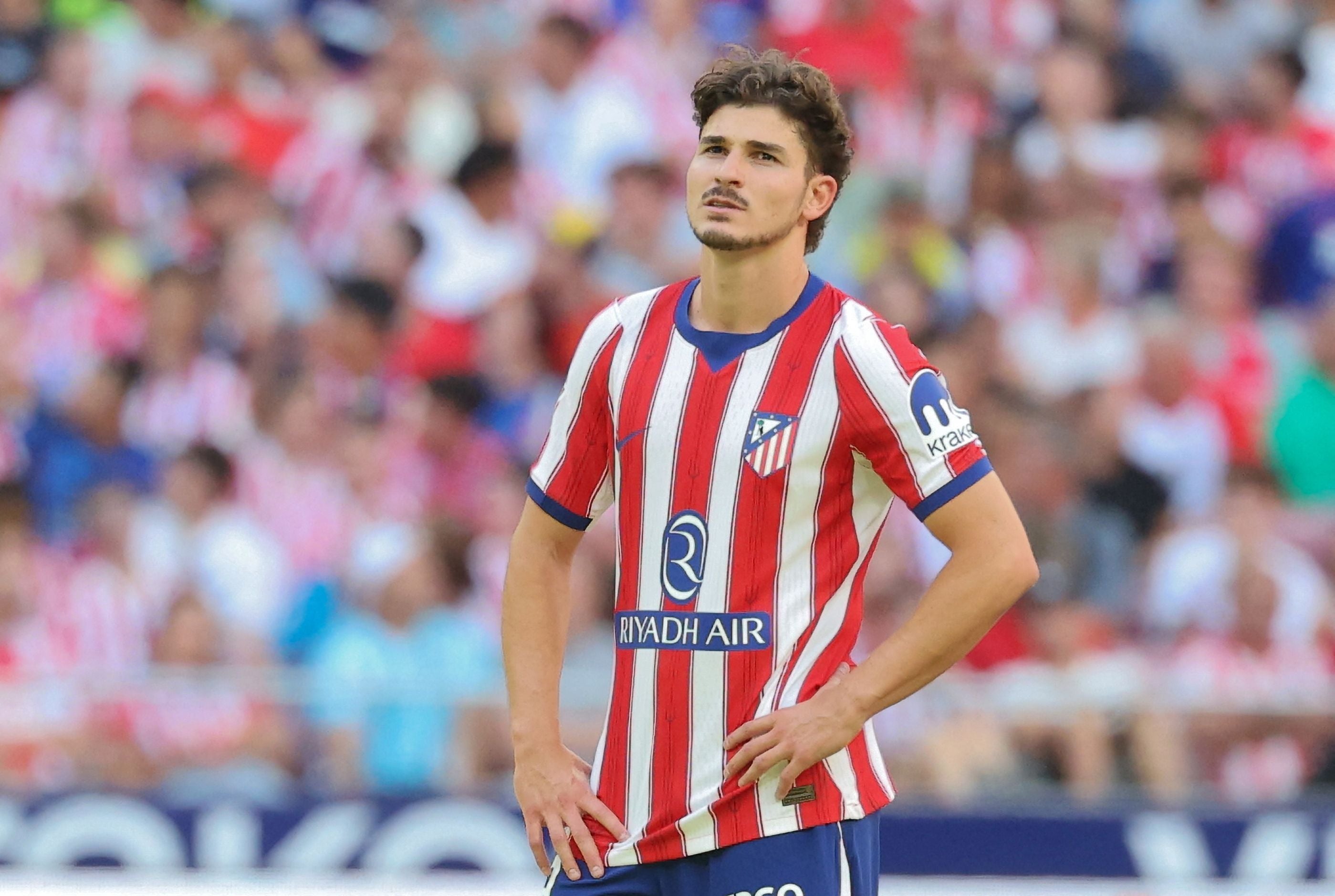 Ein Fußballspieler von Atlético Madrid steht nachdenklich auf dem Spielfeld, Zuschauer im Hintergrund.