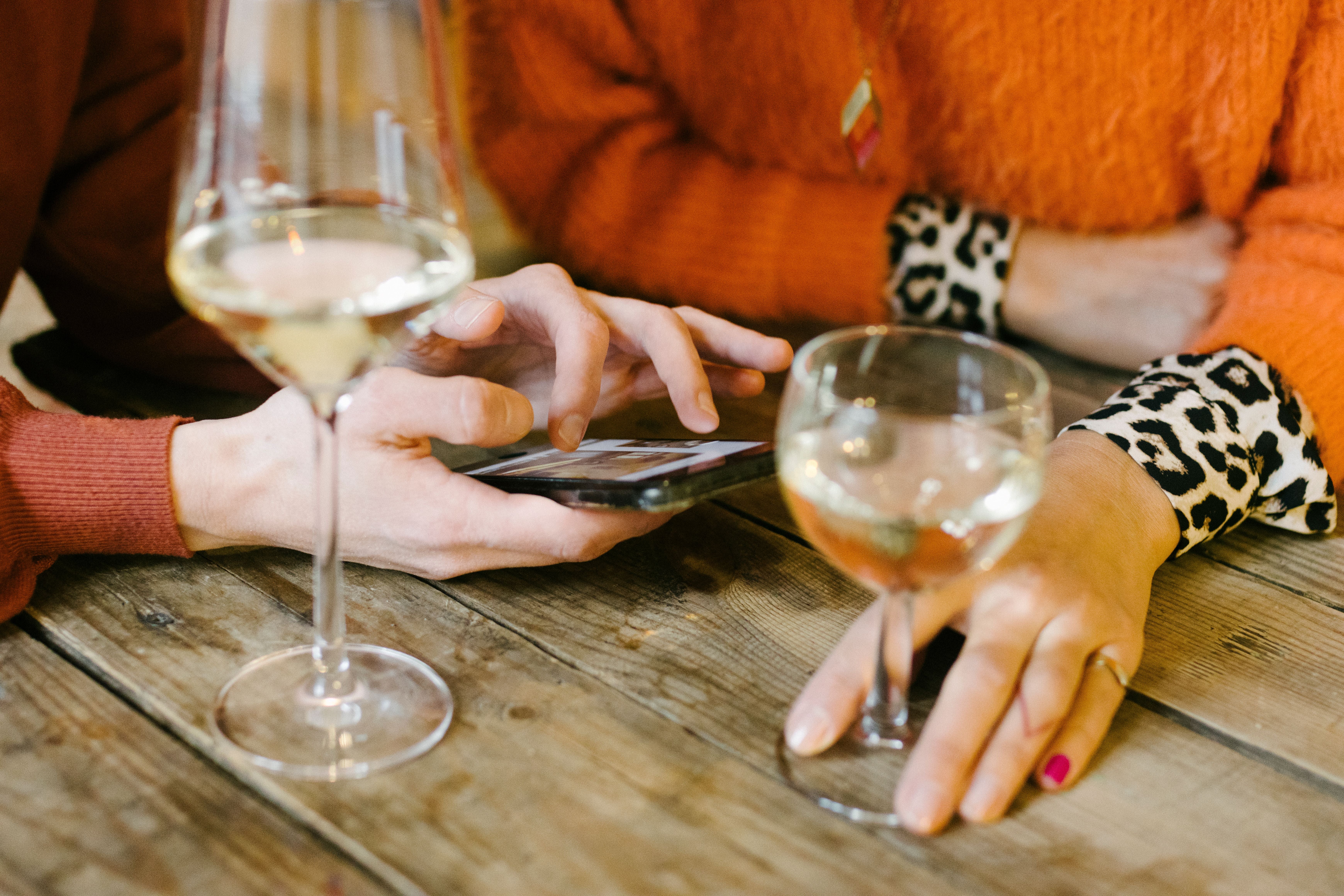 Zwei Personen trinken Weißwein und schauen gemeinsam auf ein Smartphone am Holztisch.